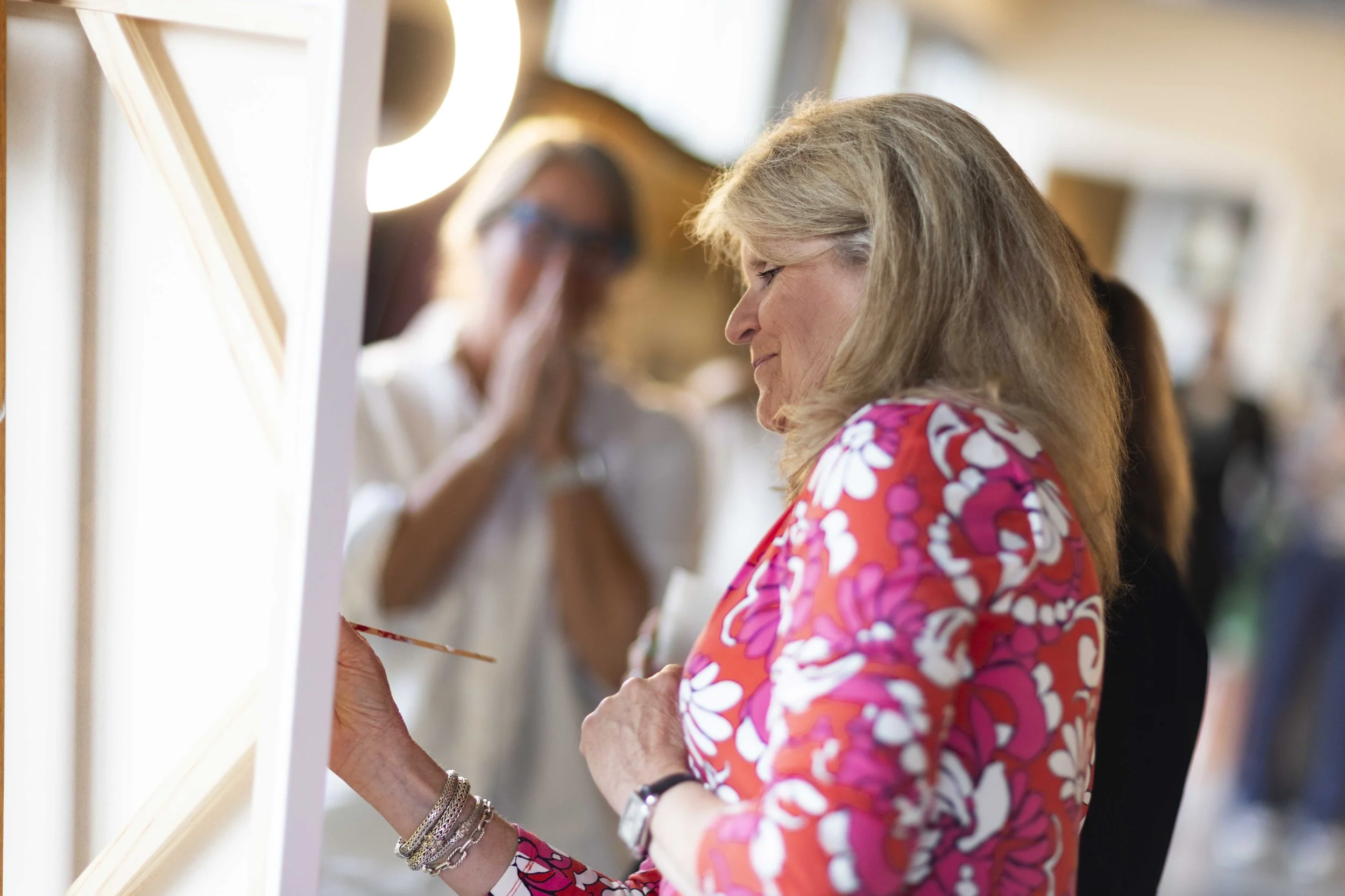 A woman with blonde hair in a floral dress is painting or drawing on a canvas, with a woman in the background clasping her hands together.