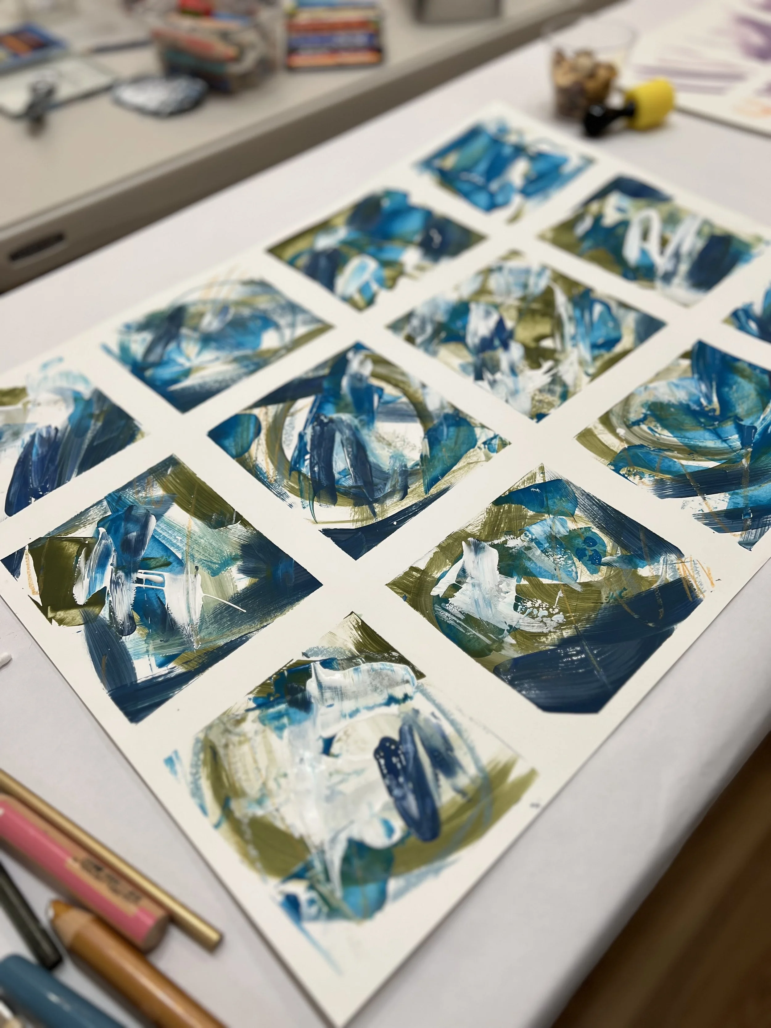 Abstract blue and green paintings on white cards arranged on a table with art supplies.