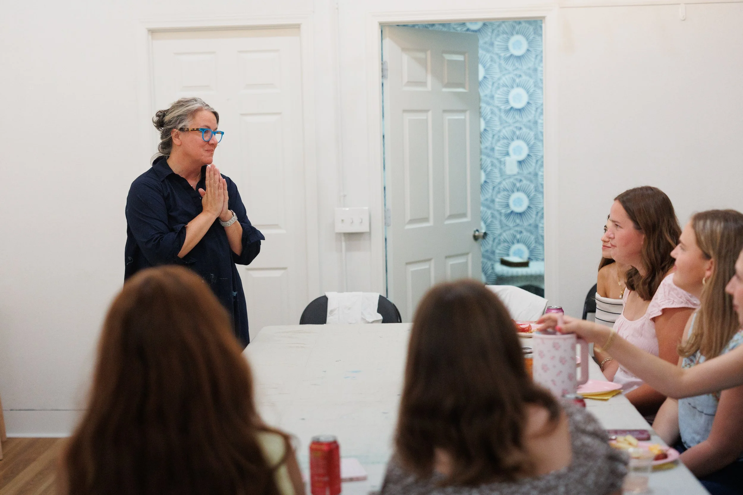 A woman stands at the head of a dining table with several women seated around it, in a room with white walls and a door leading to a room with blue floral wallpaper. The standing woman has gray hair, glasses, and is wearing a dark shirt, with hands t