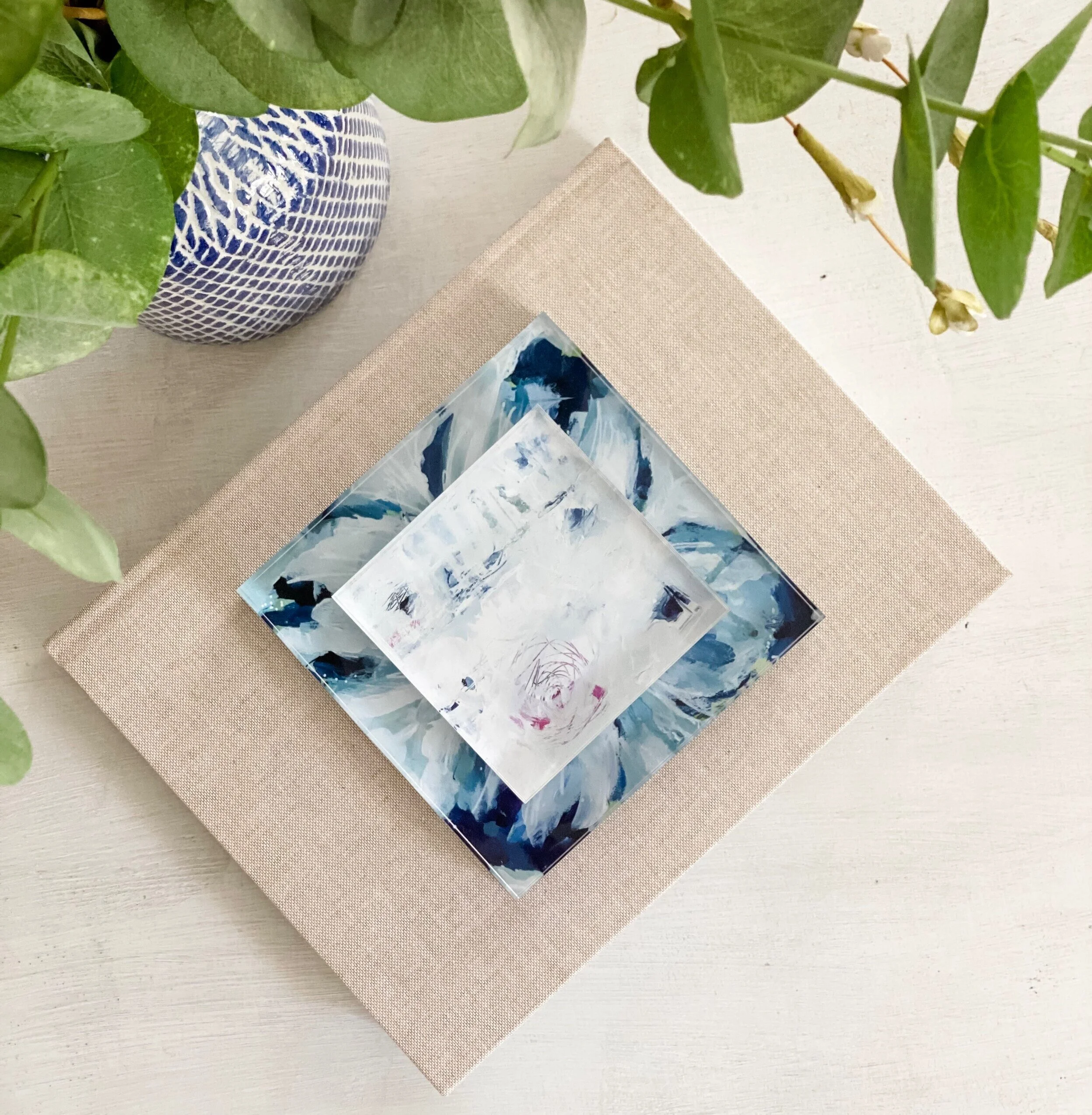 Artistic tabletop setup with a beige linen-covered book, a small abstract painting on top, and decorative green leaves and a blue-and-white patterned vase on the side.