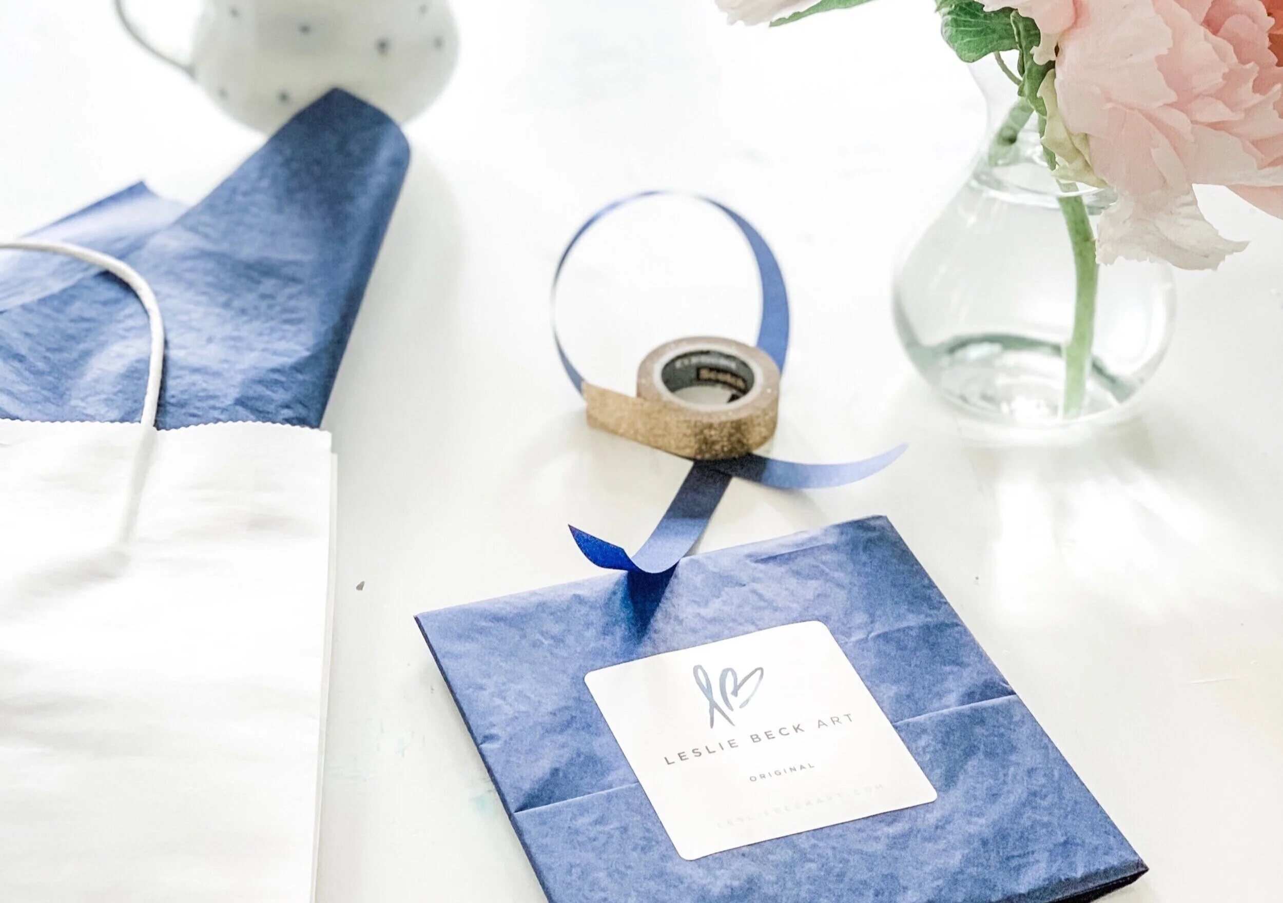 Blue wrapping paper with "Leslie Beck Art" label, gold tape, ribbon, white bag, vase with pink flowers on a table.