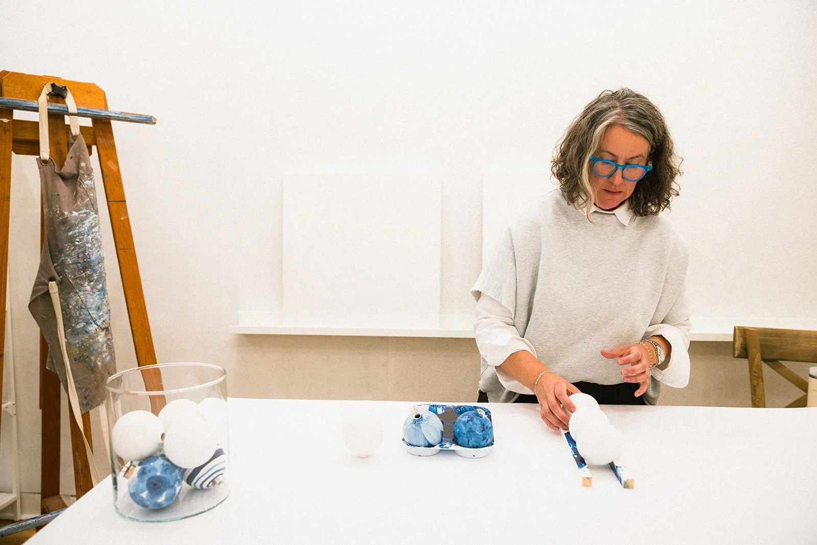 Leslie Beck working at a table with art materials including her white and blue hand painted ceramic ornaments. An easel with a paint-splattered apron hangs on the side.