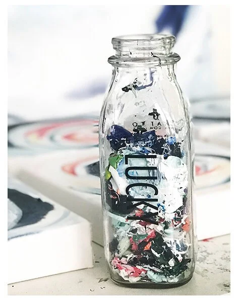Glass bottle filled with colorful paint scraps on an artist's table with canvases in the background.