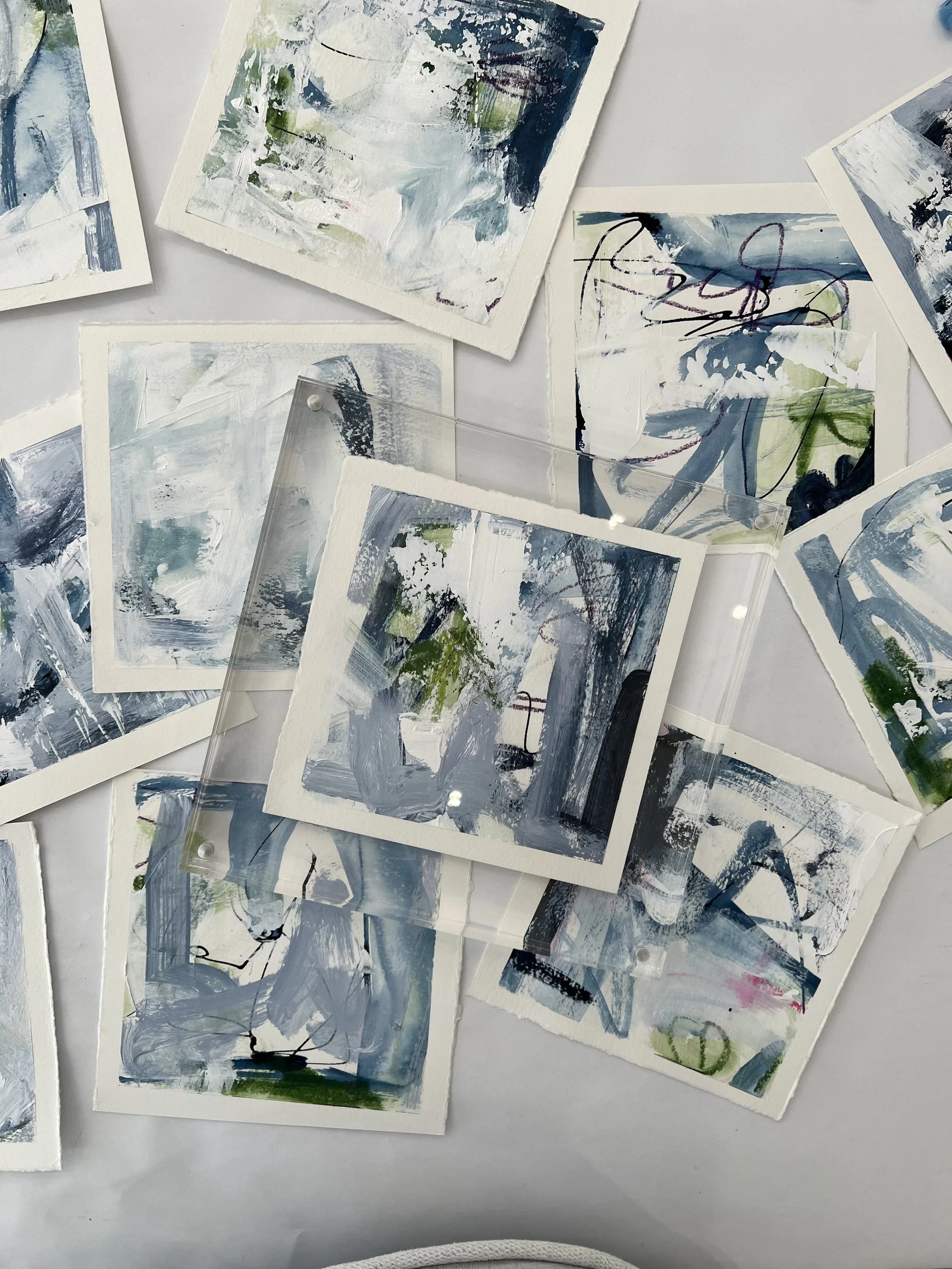 Collection of small abstract paintings on paper with various brushstrokes and colors like blue, green, white, and black, some covered with clear acrylic or glass.