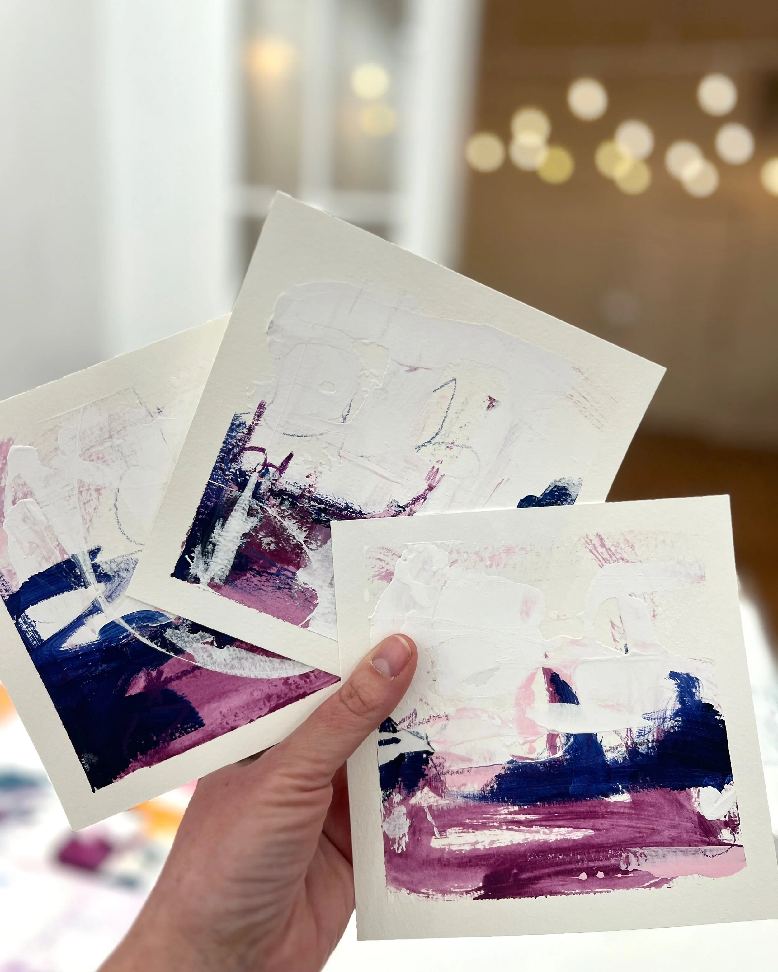 Hand holding three abstract paintings on paper with brushstrokes in pink, purple, navy blue, and white.