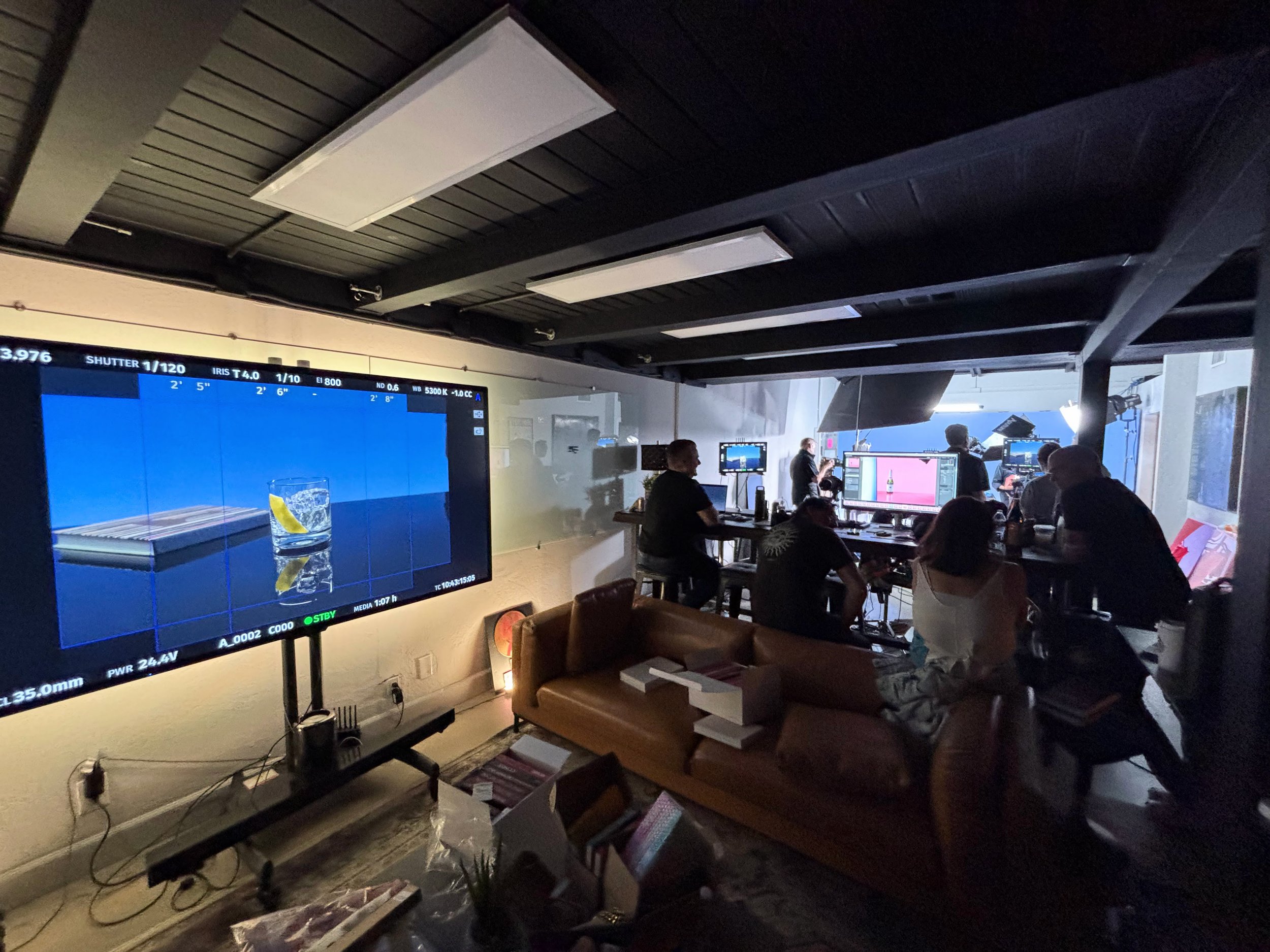 A film set or studio with people working around computers and monitors, a large screen displaying a shot of a glass with a lemon wedge, and various filming equipment in a dimly lit room.