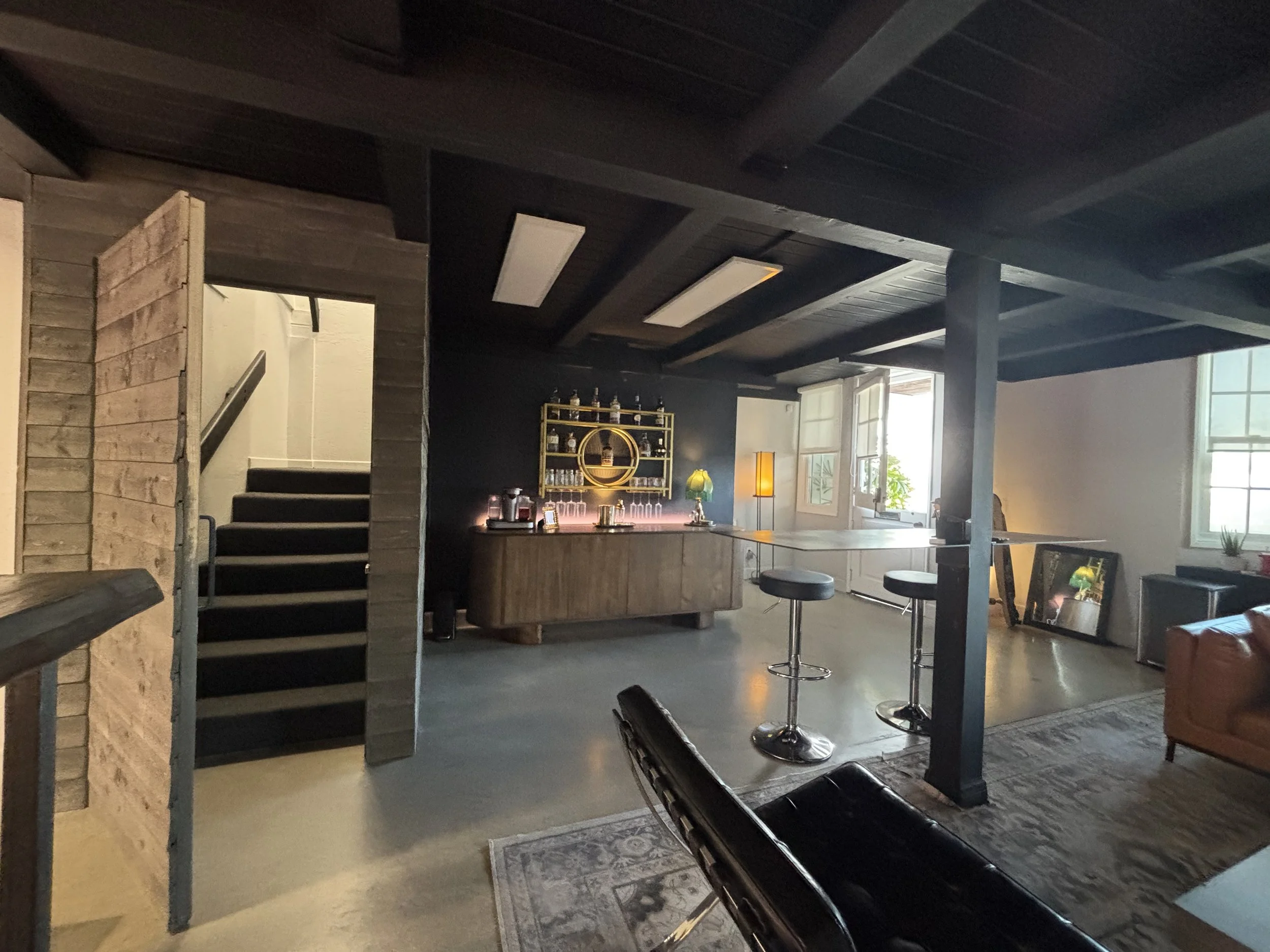 Interior of a cozy living area with a bar, high stools, and open windows, featuring dark wood accents, a wooden staircase, and modern decor.