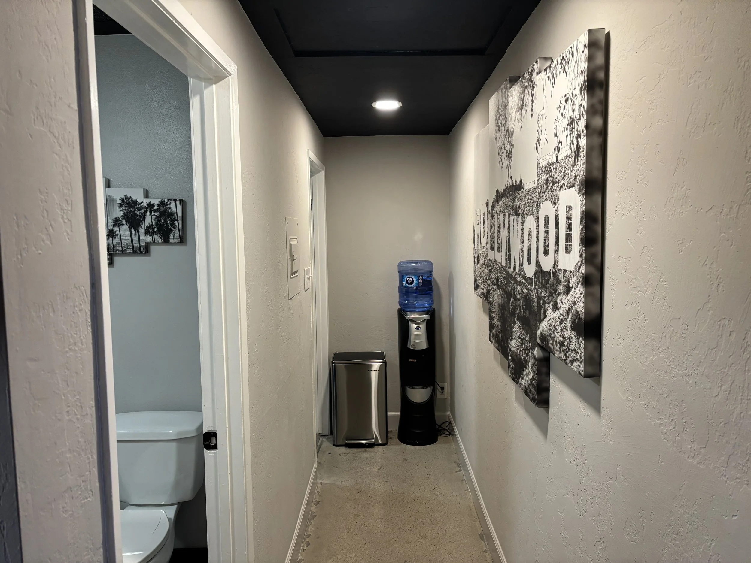 Hallway with a water cooler on the right, a trash can, and a mini-fridge. The wall on the right has a black and white artwork of the Hollywood sign. A bathroom with a toilet is visible through an open door on the left.