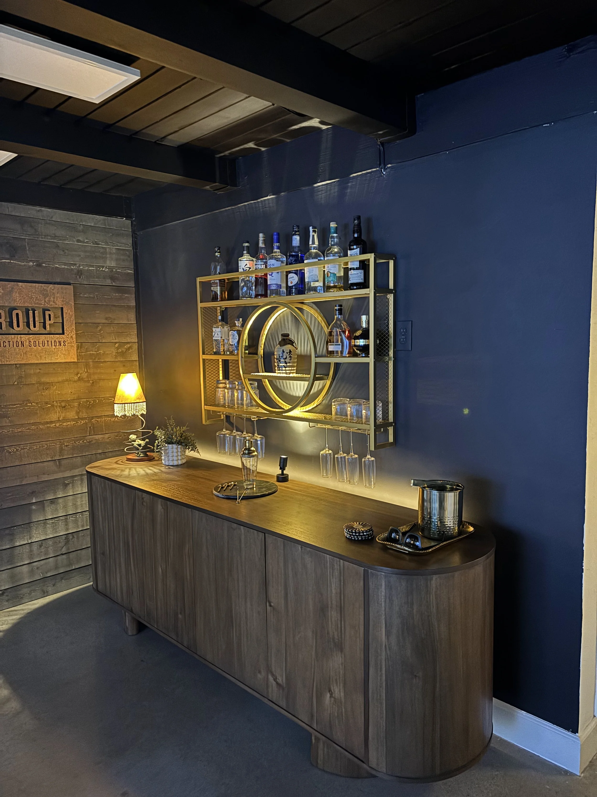 A home bar setup with a wooden cabinet, a decorative wall shelf with liquor bottles and wine glasses, a small lamp, and some decorative items.