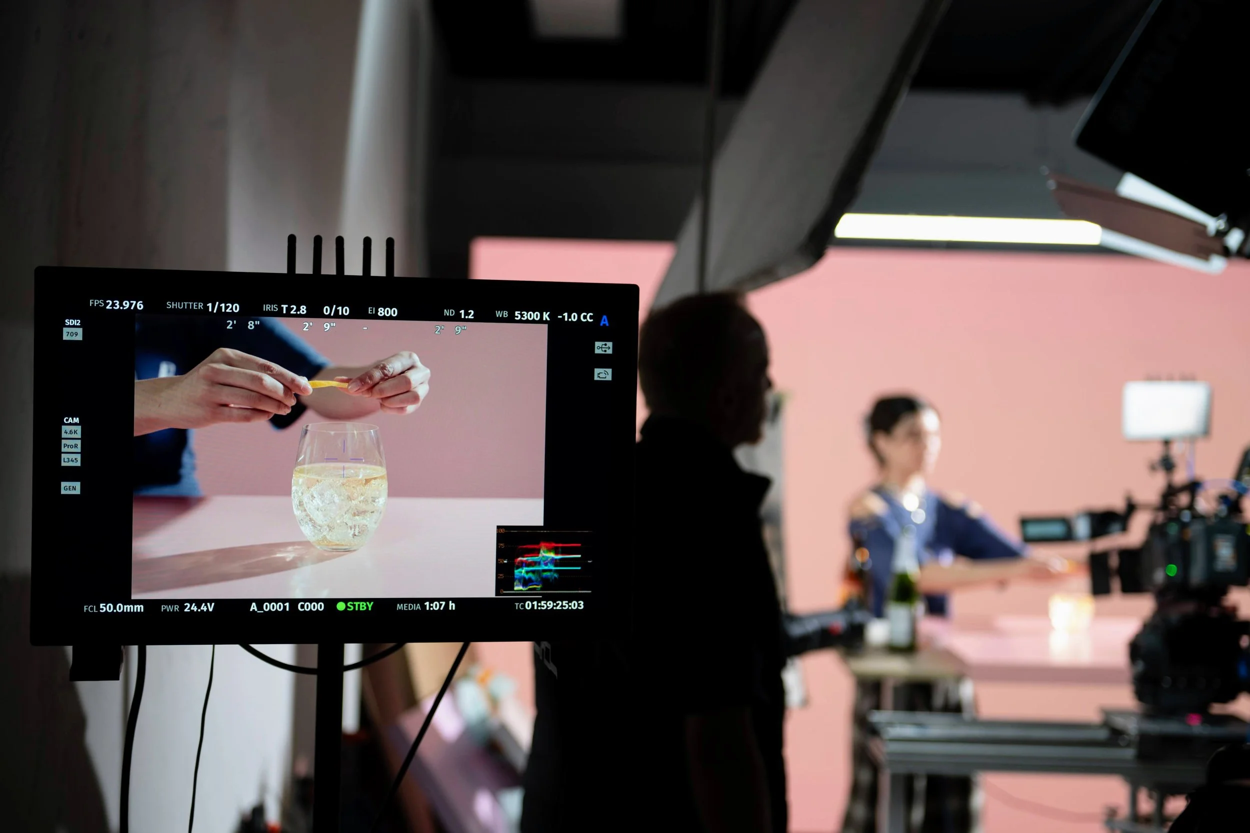 Behind-the-scenes of a video shoot with a monitor displaying a close-up of hands placing a lemon peel into a glass of lemonade on a pink background, with crew members and filming equipment visible in a dim studio.
