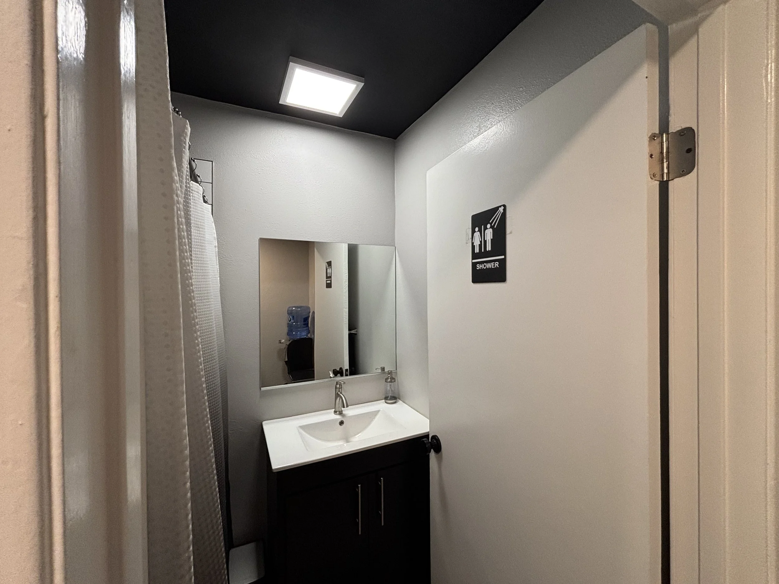 Small restroom with a sink, mirror, shower sign on the door, water cooler in the background, and a ceiling light.