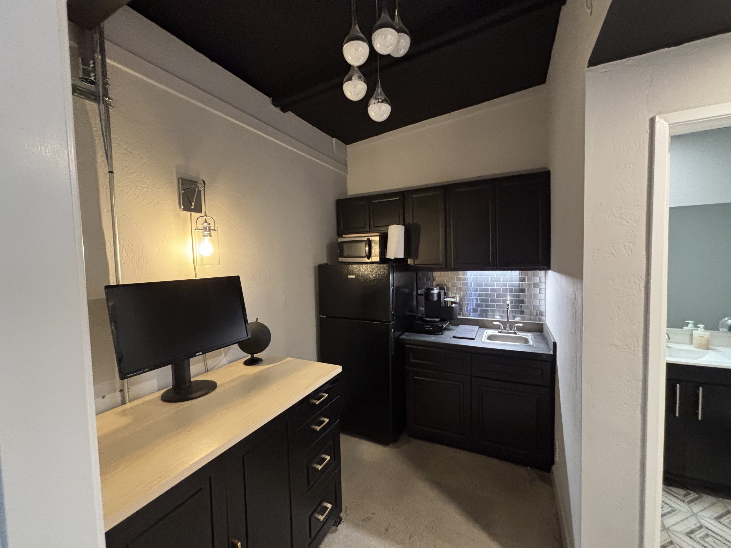 Small kitchen area with black refrigerator, black cabinets, a microwave, coffee maker, stainless steel sink, and a tile backsplash. Adjacent workspace with a monitor and a lamp.