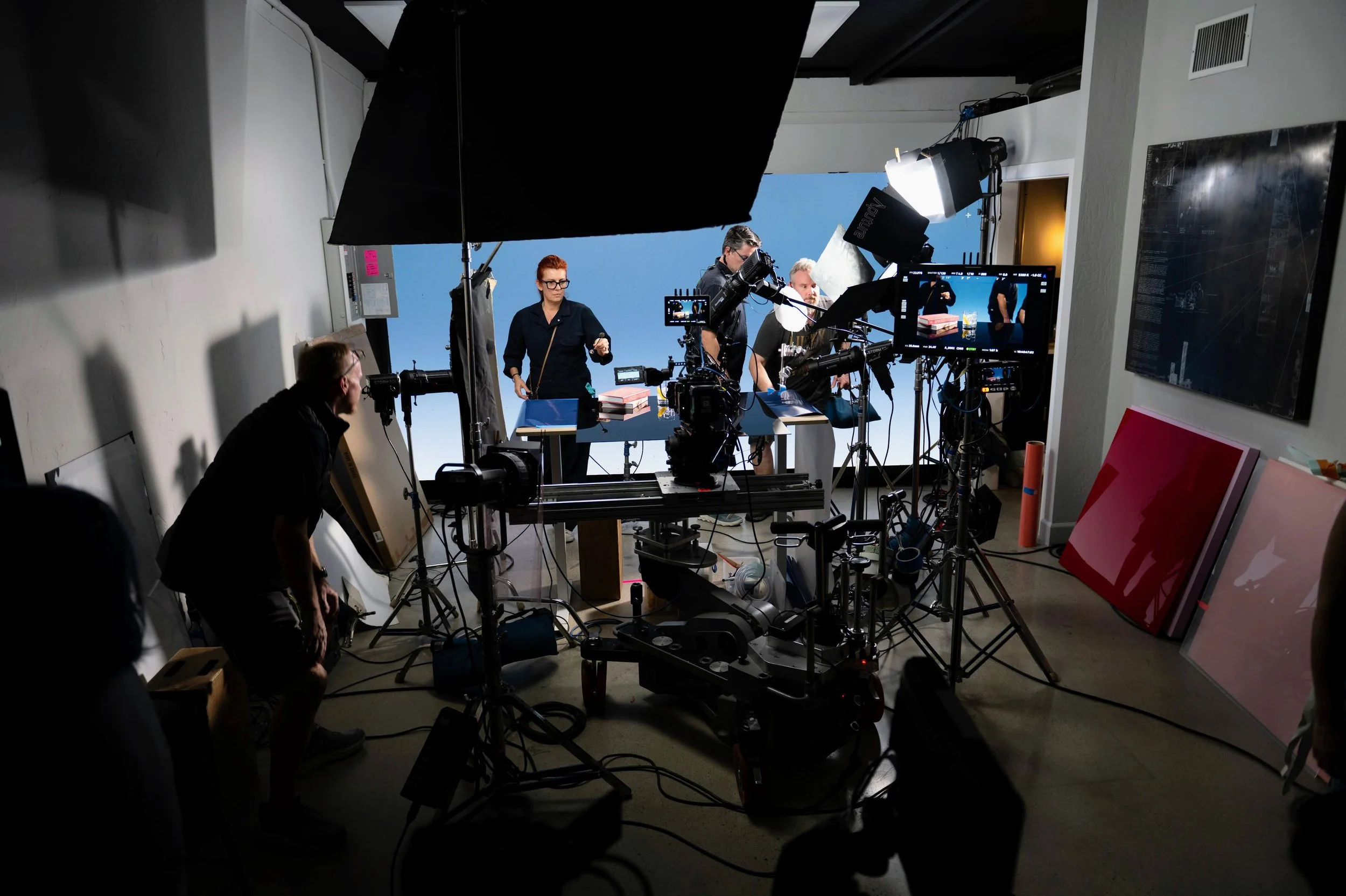 A behind-the-scenes photo of a film or photo shoot setup in a studio, with multiple lights, cameras, and crew members working around a table with books and props, a blue backdrop, and various equipment and set pieces.