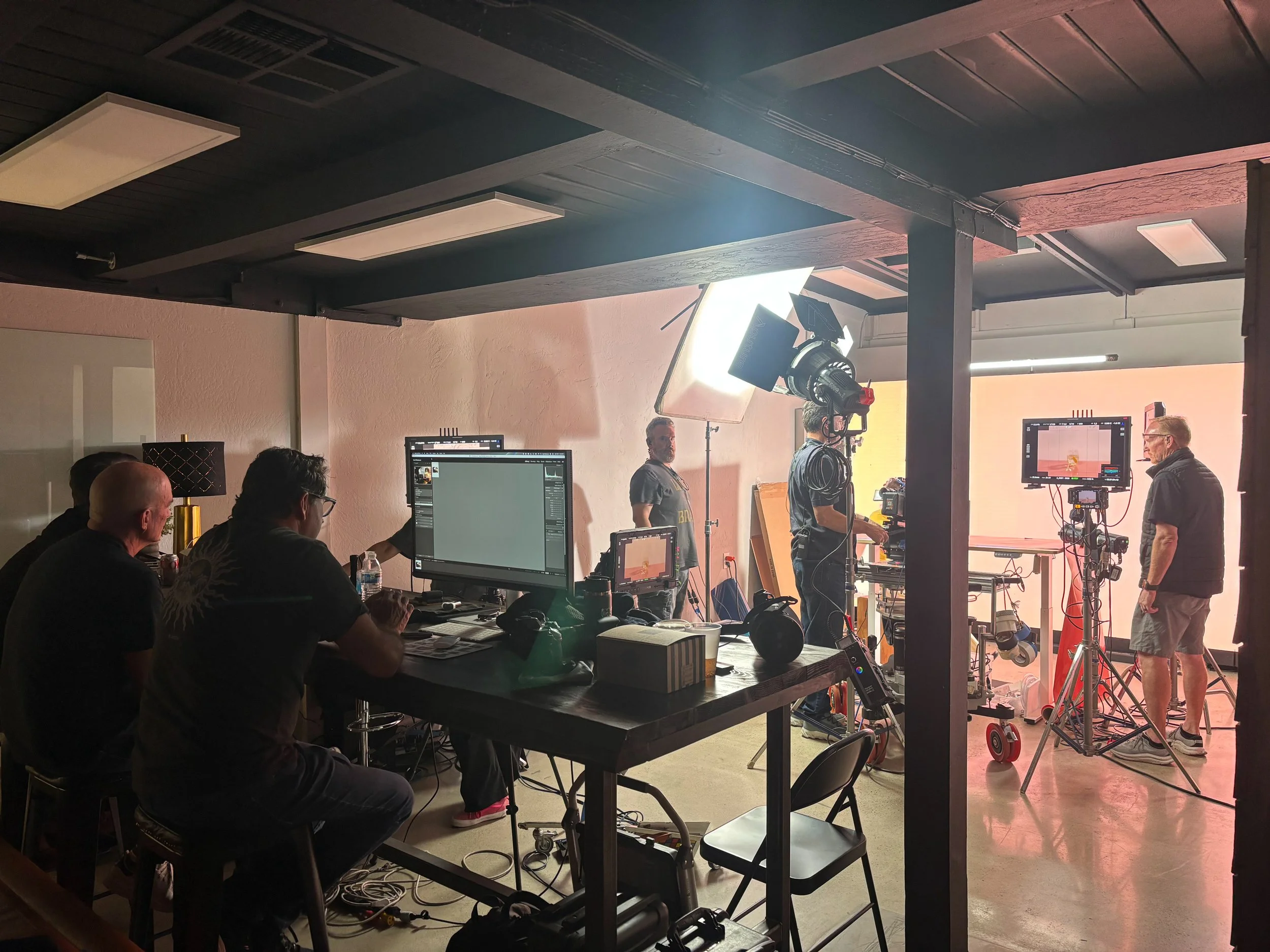 Film production crew working on a set with lighting and camera equipment, with crew members seated at a table monitoring screens and others setting up in front of a green screen.