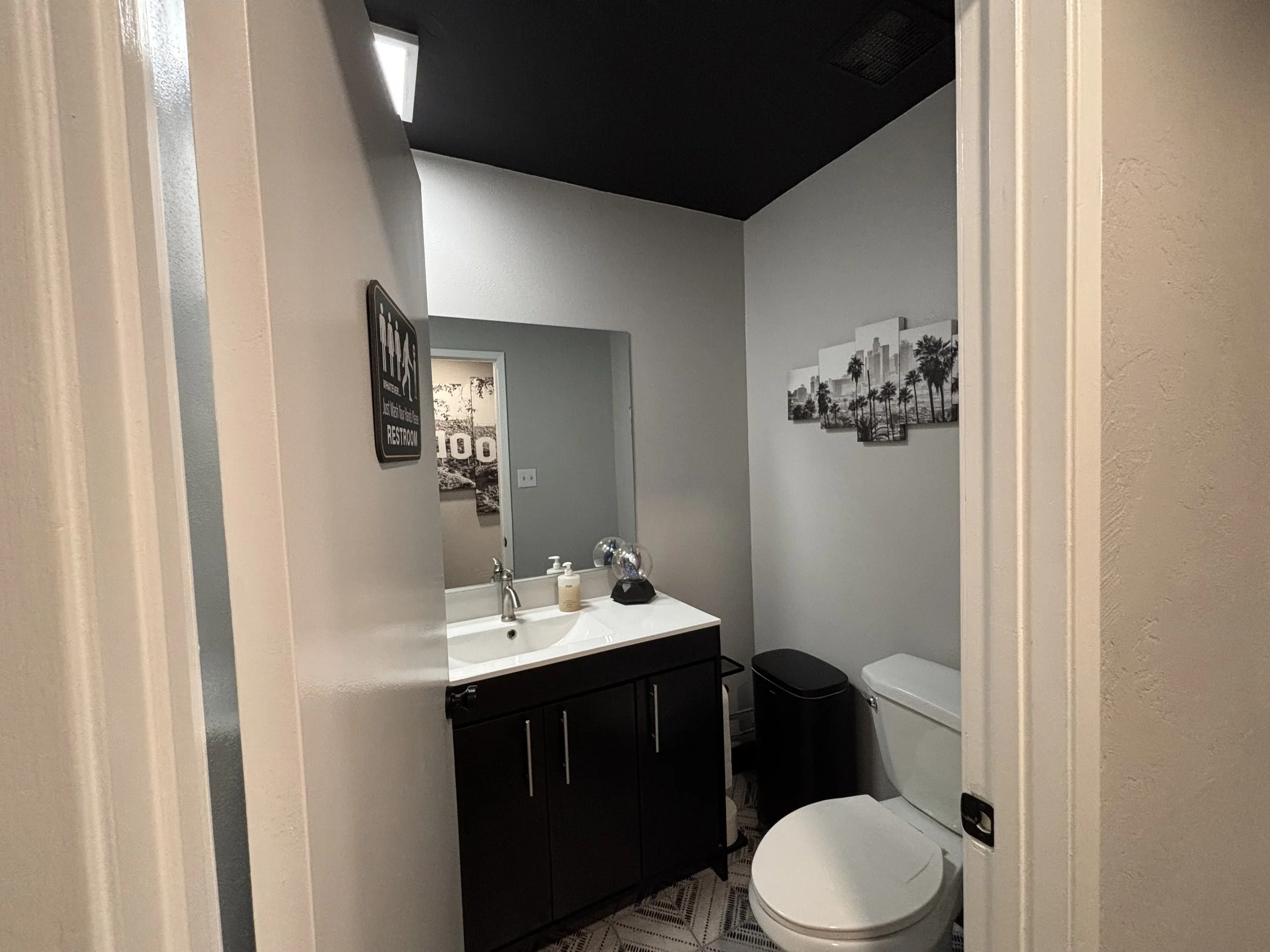 A modern bathroom with a black vanity, white sink, mirror, black trash bin, toilet, and wall art of palm trees and cityscape. Decorative items and soap on the sink counter.