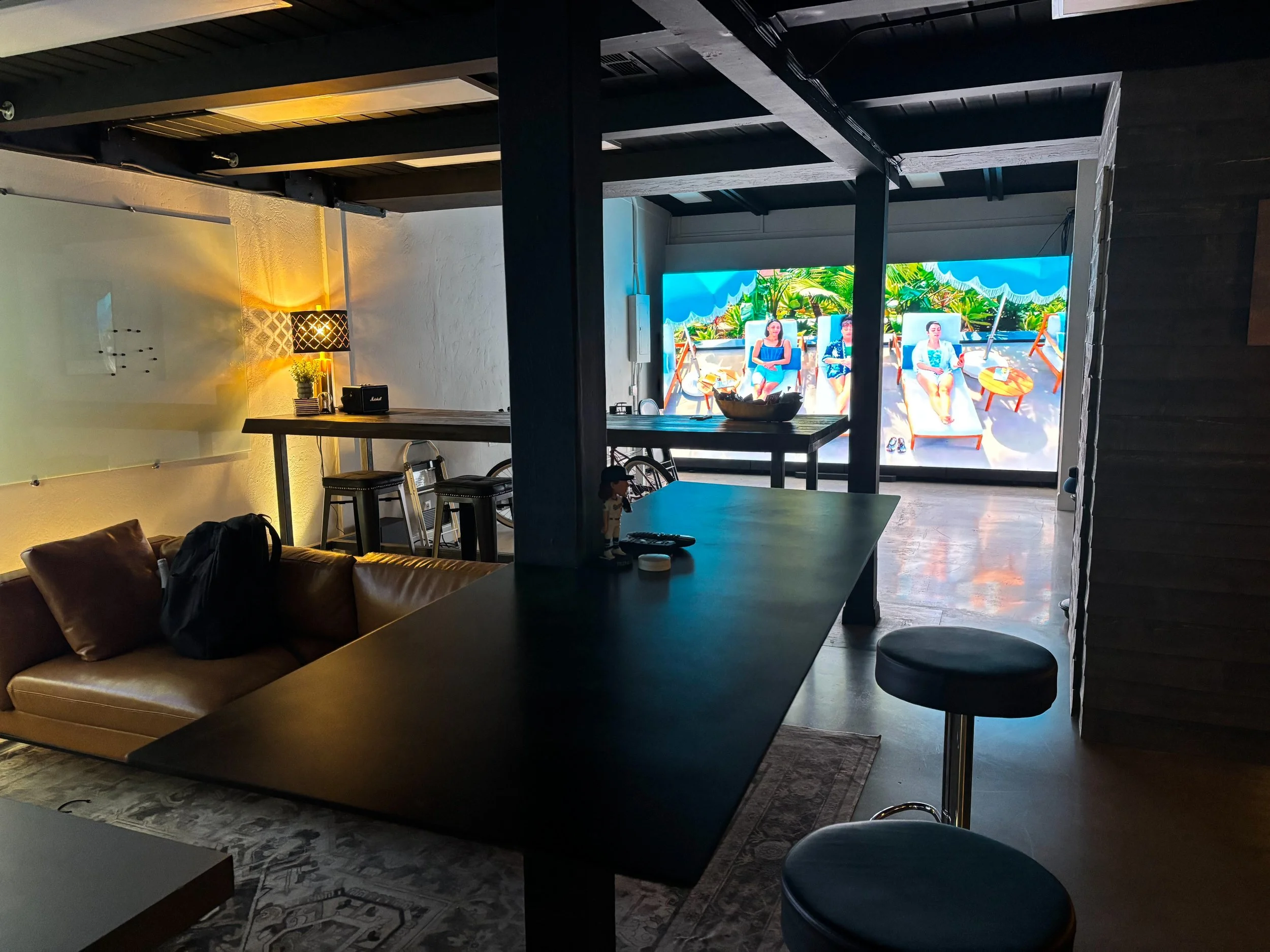 Indoor lounge area with a couch, bar-height table, and snacks. Large screen TV displays people relaxing by a pool under umbrellas.