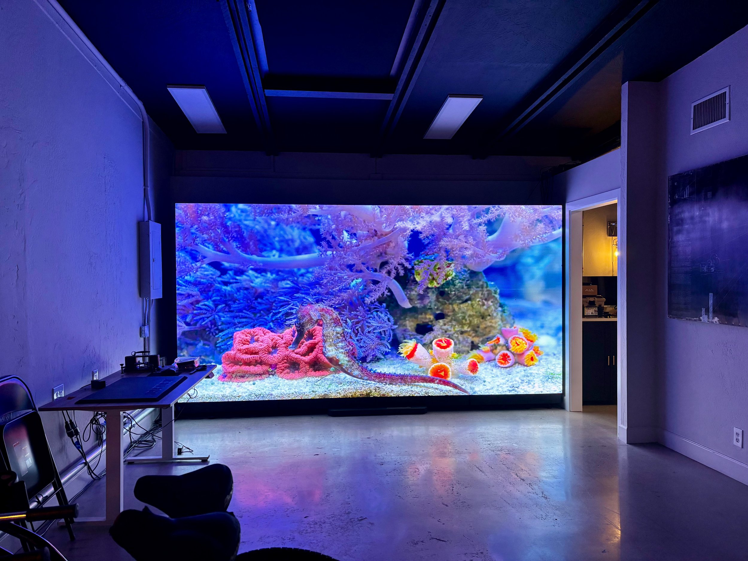 Large digital screen displaying an underwater scene with coral, fish, and sea creatures in a room with minimal furniture and purple lighting.
