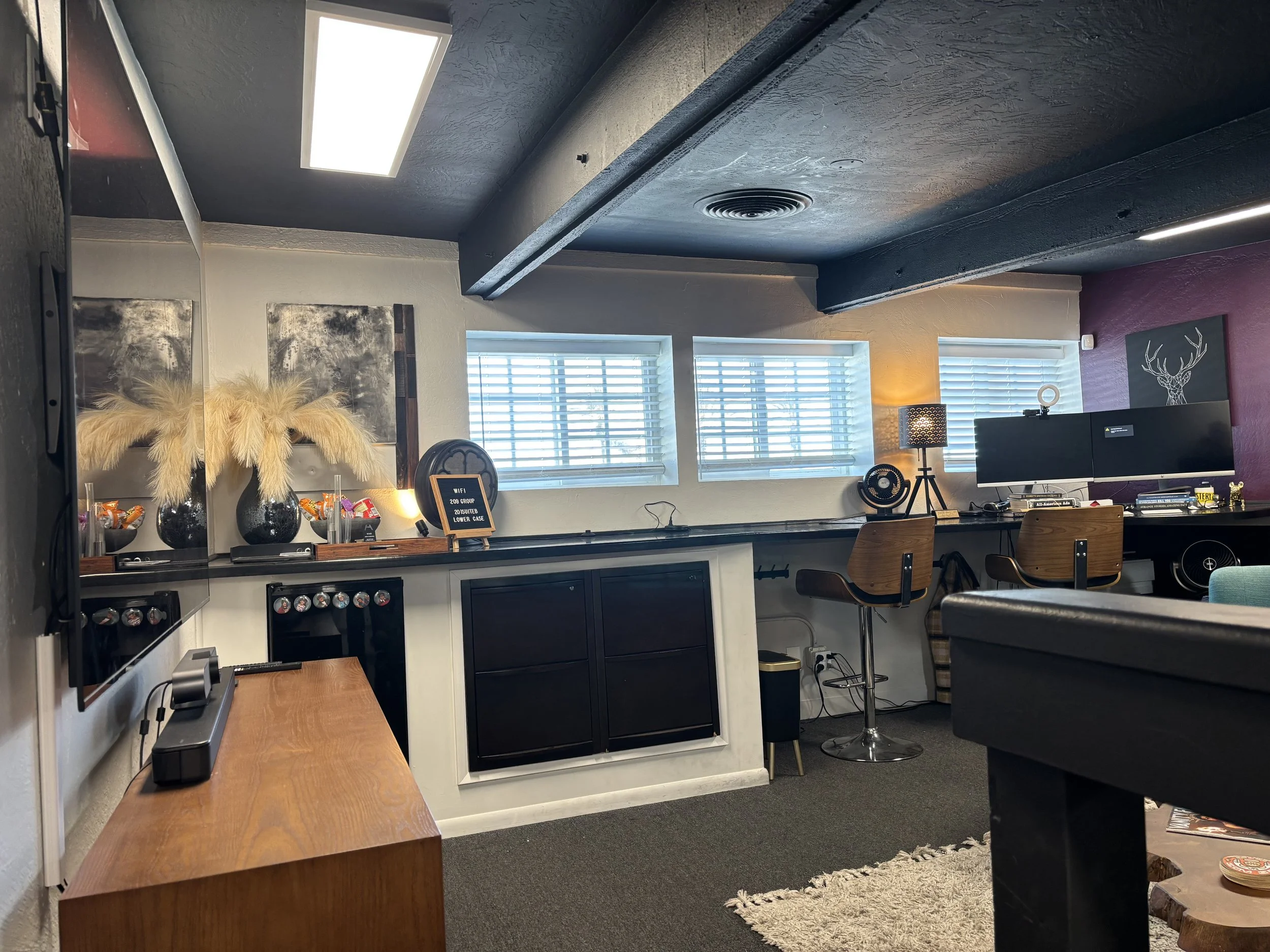 An office space with a wall-mounted flat-screen monitor, wooden chairs, a desk lamp, decorative artwork, and large windows with blinds letting in natural light.