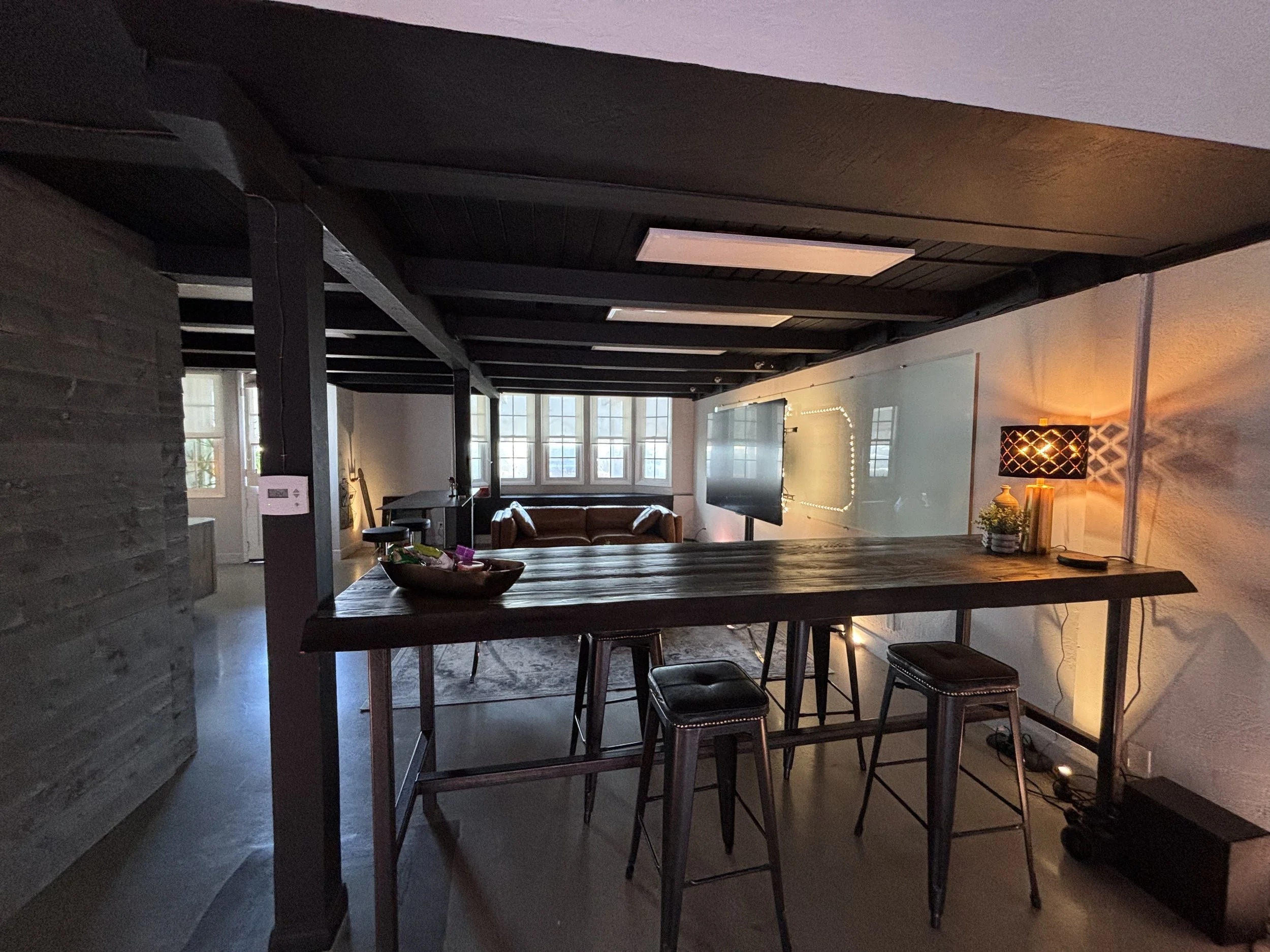 Interior of a cozy living room with a large window, a brown leather sofa, flat-screen TV, small table with plants, and a high table with three bar stools, warm lighting, and decorative items.