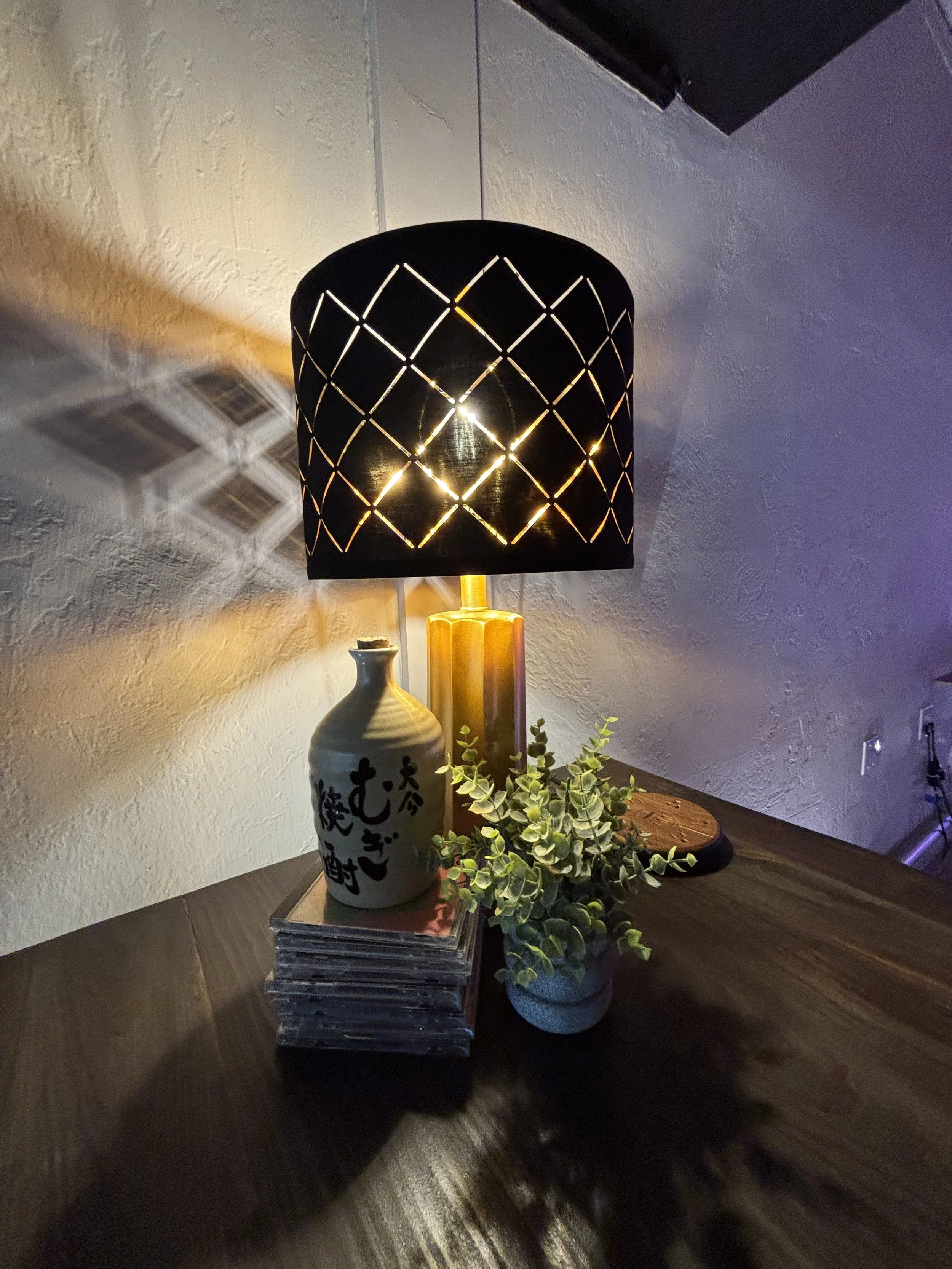 A black table lamp with a diamond-shaped cutout pattern casting light and shadows on the white wall behind it. The lamp sits on a dark wooden surface alongside a decorative ceramic vase with Japanese characters, a small potted plant, and a stack of m