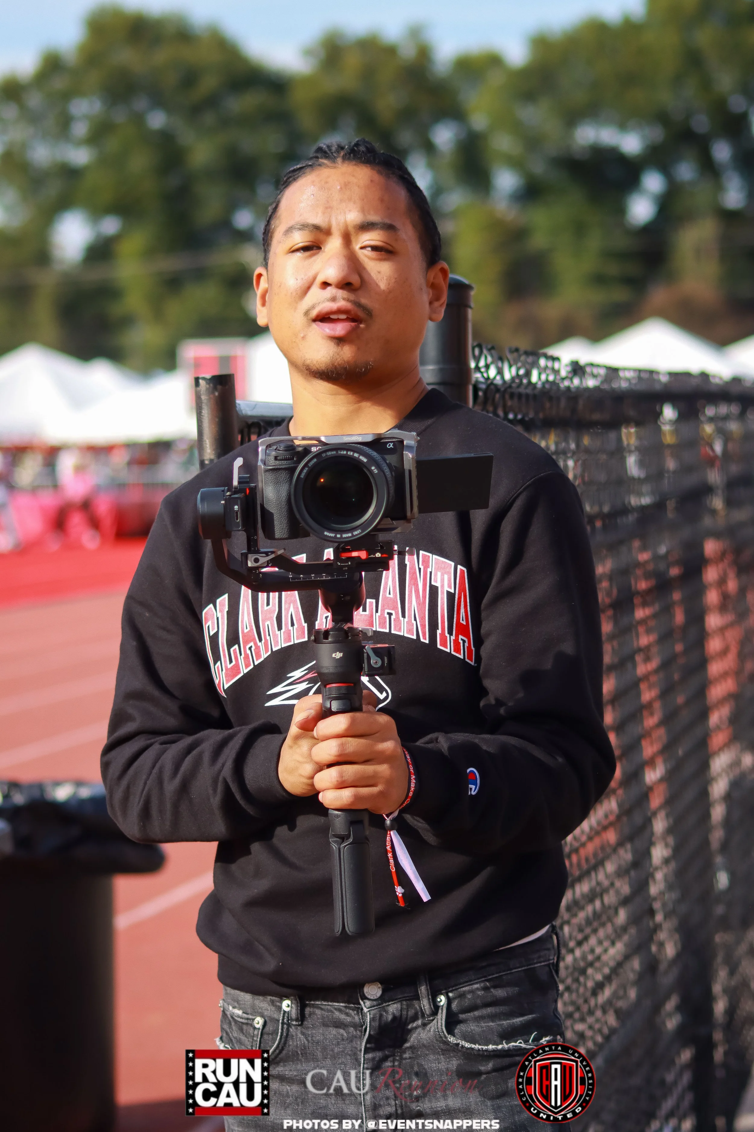 A young man holding a camera on a tripod at an outdoor event, with a chain-link fence and trees in the background. He is wearing a black hoodie with red and white text and has a serious expression.