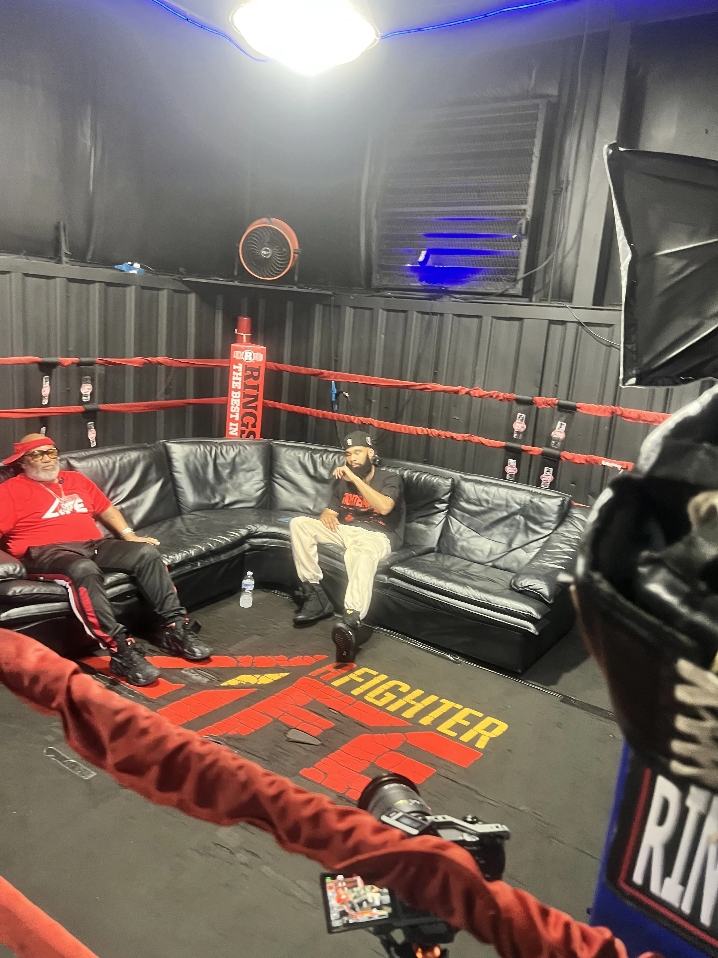 People sitting on a black leather couch inside a boxing gym, which has a red and black boxing ring. One person is wearing a red shirt and hat, and another is in a black shirt and beige pants. The gym has gray walls, a large bright light on the ceiling, and various equipment including a fan, a vent, and a blue-lit fixture. The floor has the words "FIGHTER" and other text in yellow and red.
