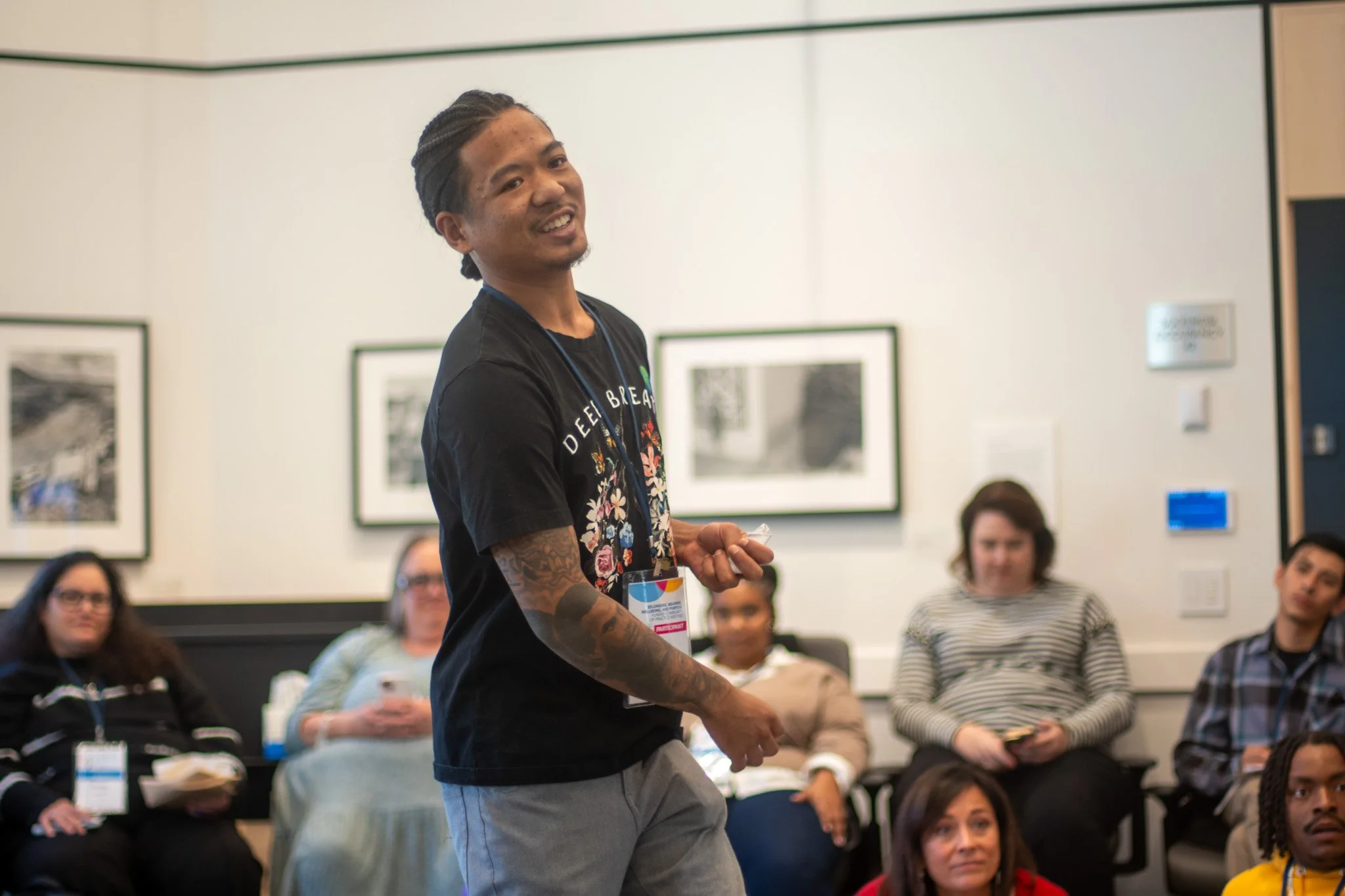 A man with braided hair and tattoos on his arms standing and speaking in front of a seated group of diverse people in a conference room with framed pictures on the wall.