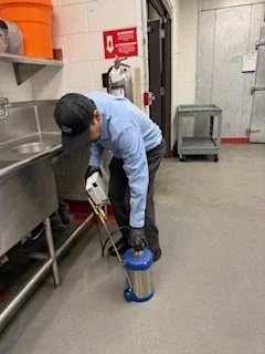 Person wearing a blue shirt and black cap using a mop and bucket in an industrial or commercial kitchen.