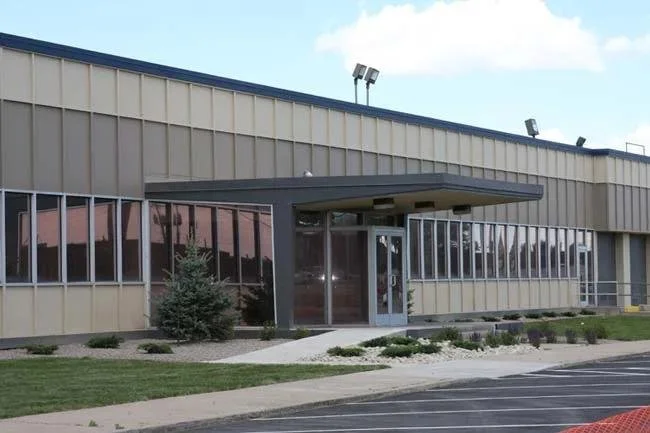 Exterior view of a commercial building with large windows, a glass sliding door entrance, a small landscaped area with shrubs, and an empty parking lot in front.