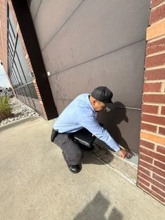 A pest control techinican crouching near a brick and gray wall, measuring or inspecting the sidewalk.