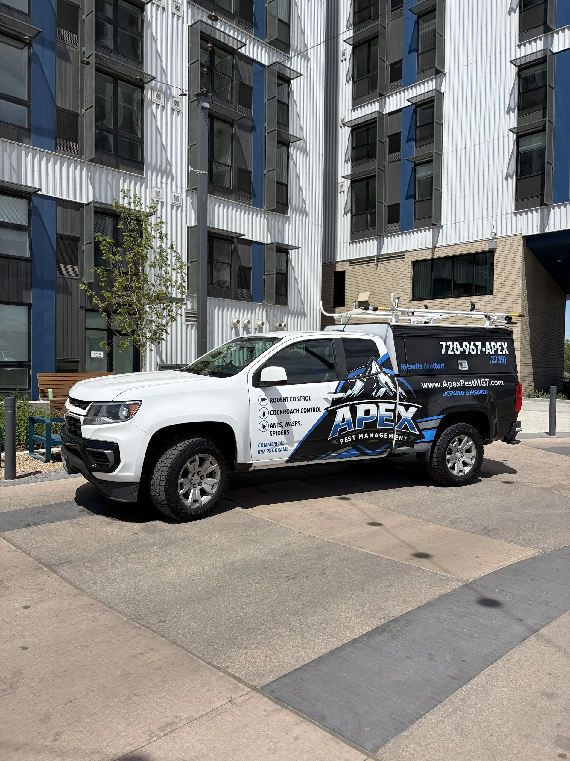 A white pest control truck with the logo and contact information parked in front of a modern apartment building with blue, white, and yellow-brick exterior walls. The truck advertises pest control services for rodents, cockroaches, ants, wasps, and spiders.
