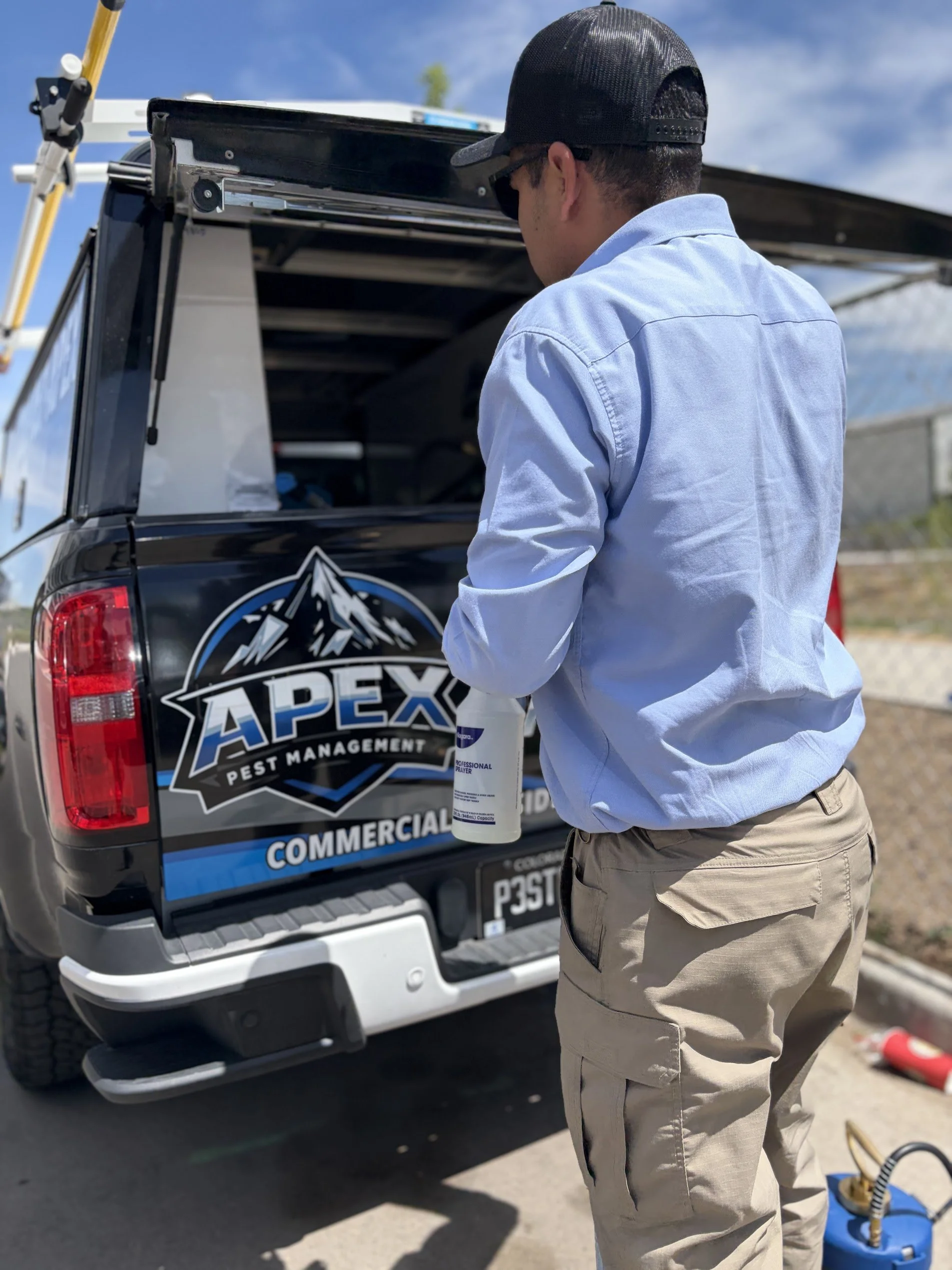 A man wearing a black cap, sunglasses, a light blue shirt, and khaki pants standing next to a black truck with an Apex Pest Management logo, holding a bottle of pest control spray.