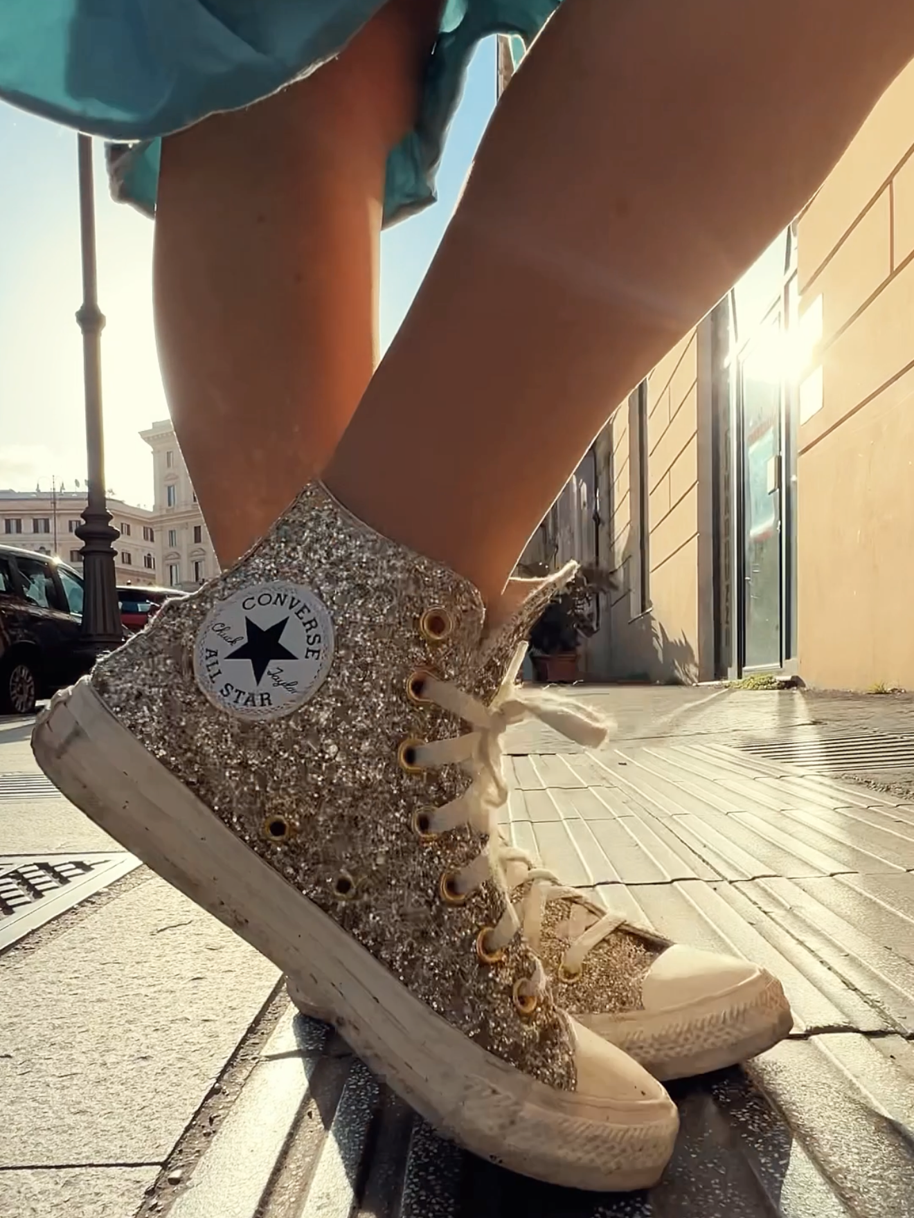 Close-up of a person wearing glittery Converse All Star sneakers on a city sidewalk during sunset, with the sun shining through a doorway.
