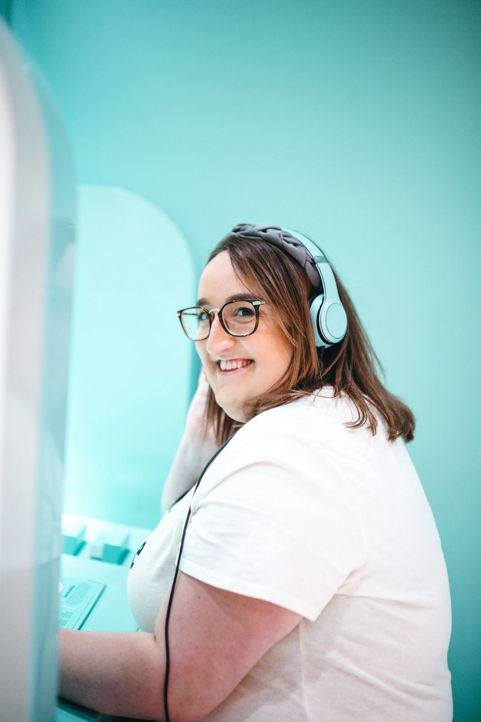 A woman with glasses and brown hair wearing a headset, smiling while sitting at a table with a turquoise wall in the background.