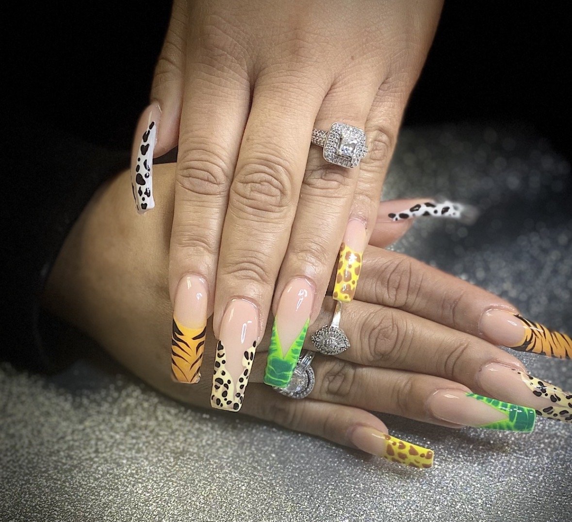 Hands with colorful animal print nails and diamond rings, resting on a sparkly surface.