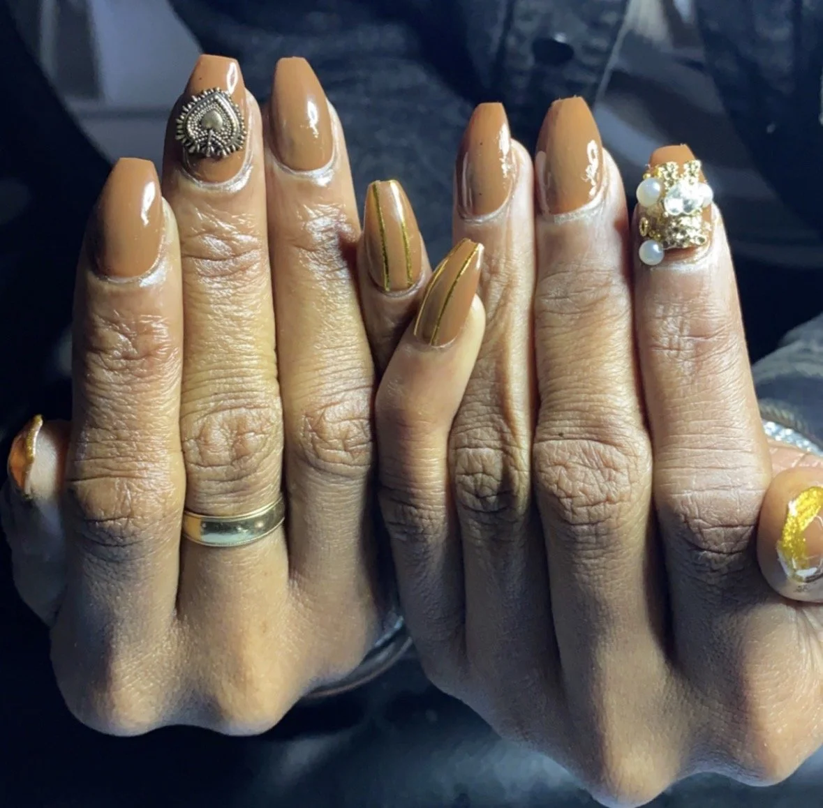 Hands with manicured nails painted in brown and gold, adorned with rings and decorative nail art, including pearls and gold accents.