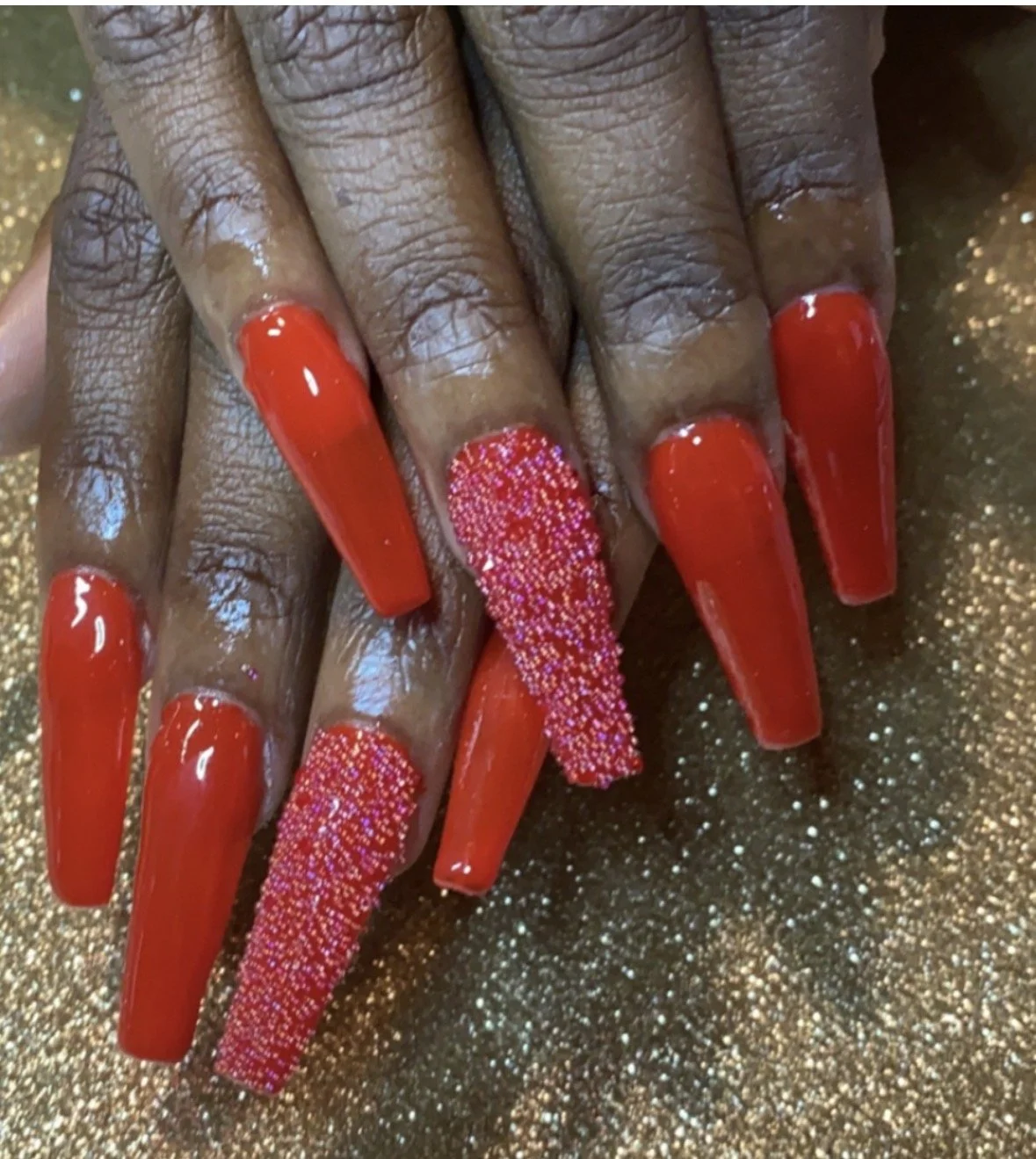 Close-up of a hand with long, square-shaped red acrylic nails, two of which feature pink and white glitter accents, resting on a glittery gold surface.