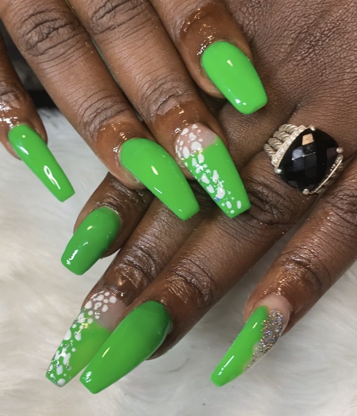 Nail art featuring bright green nail polish with white accents, glitter, and bubble designs, along with a large black gemstone ring.