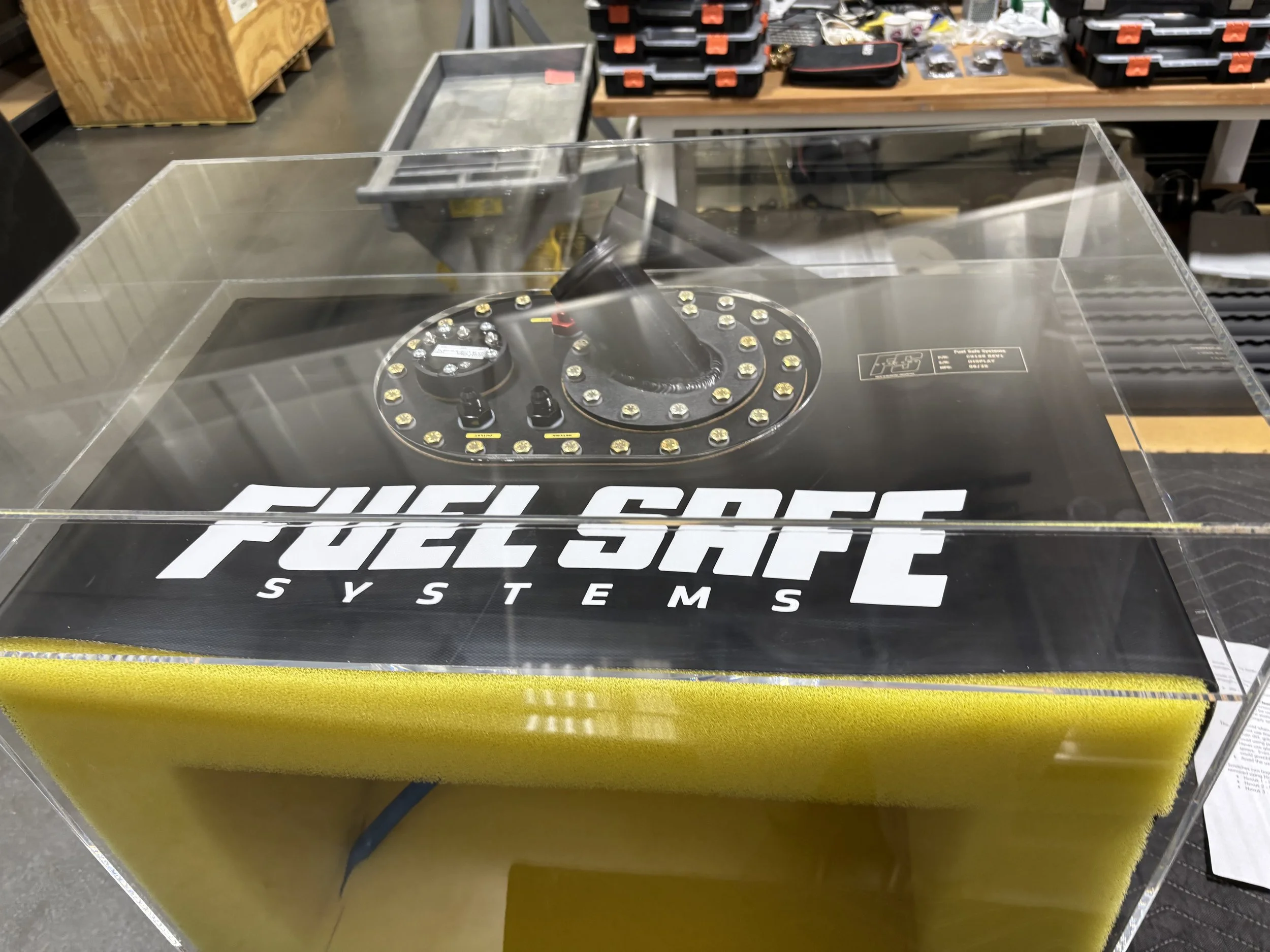 A Fuel Safe Systems racing fuel cell with a black top, equipped with a vent valve and fuel outlet, enclosed in a transparent protective cover, with a yellow foam padding at the bottom, placed on a workbench in a workshop.