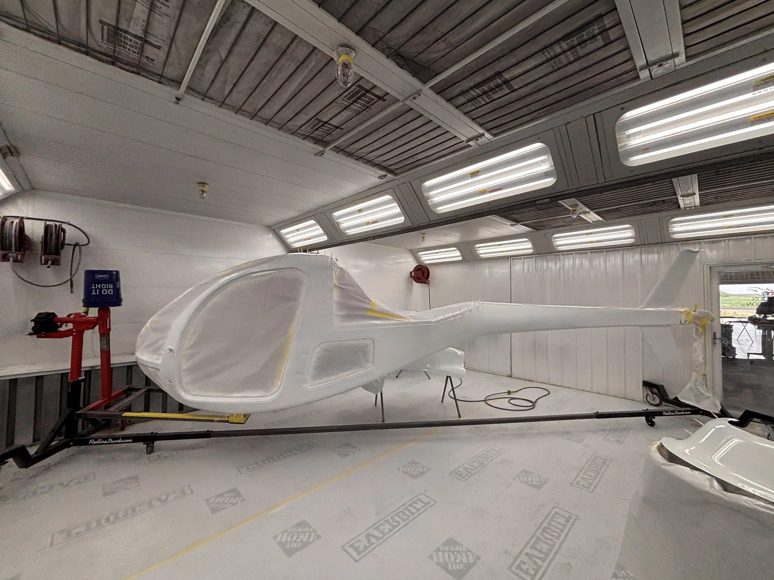 A white helicopter fuselage in a workshop or hangar, with parts covered and masking tape, surrounded by tools and equipment.