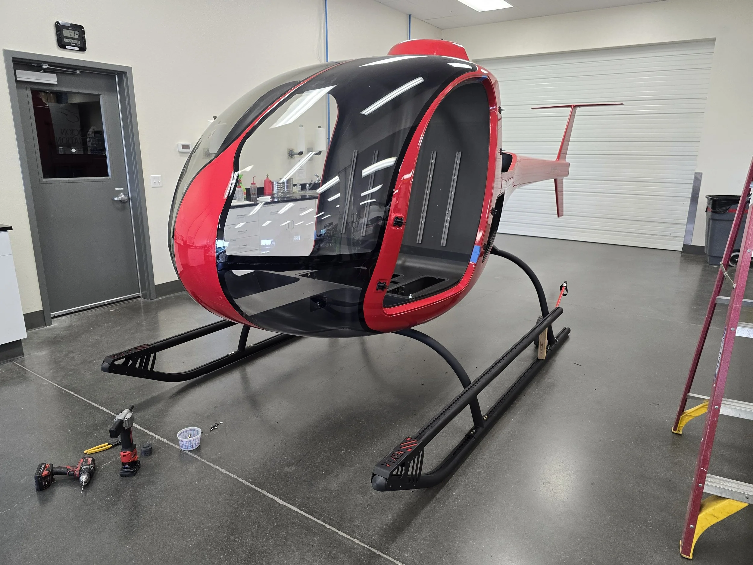 Red and black personal helicopter inside a workshop, with tools and ladders nearby.