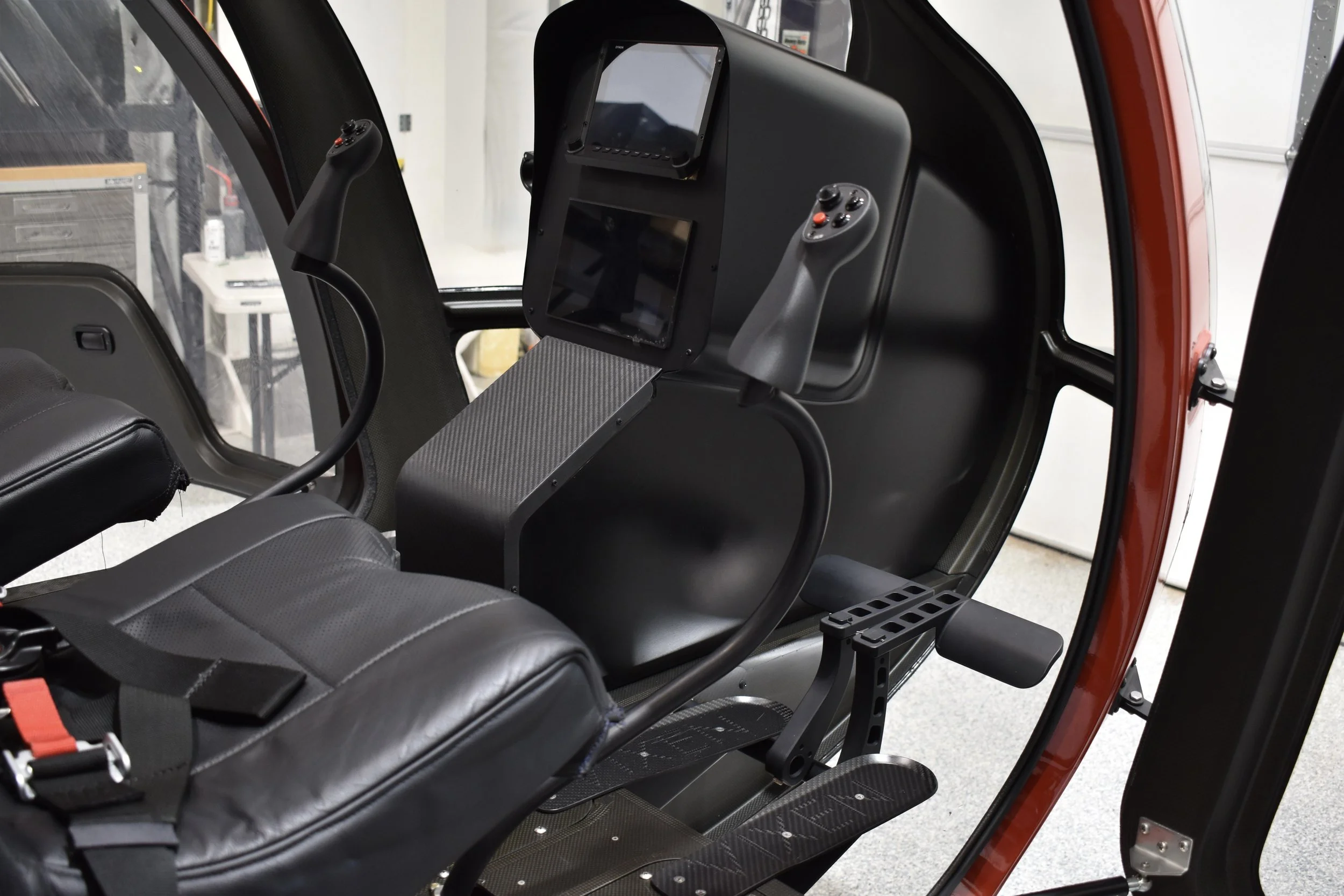 Interior view of a helicopter cockpit with a black seat, control sticks, and digital displays.