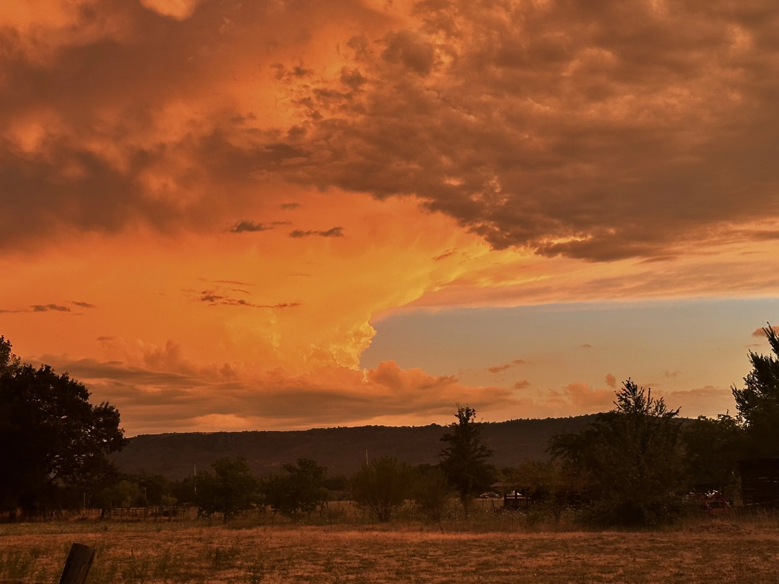 A sunset or sunrise sky with large, orange and dark clouds over a rural landscape with trees and hills.