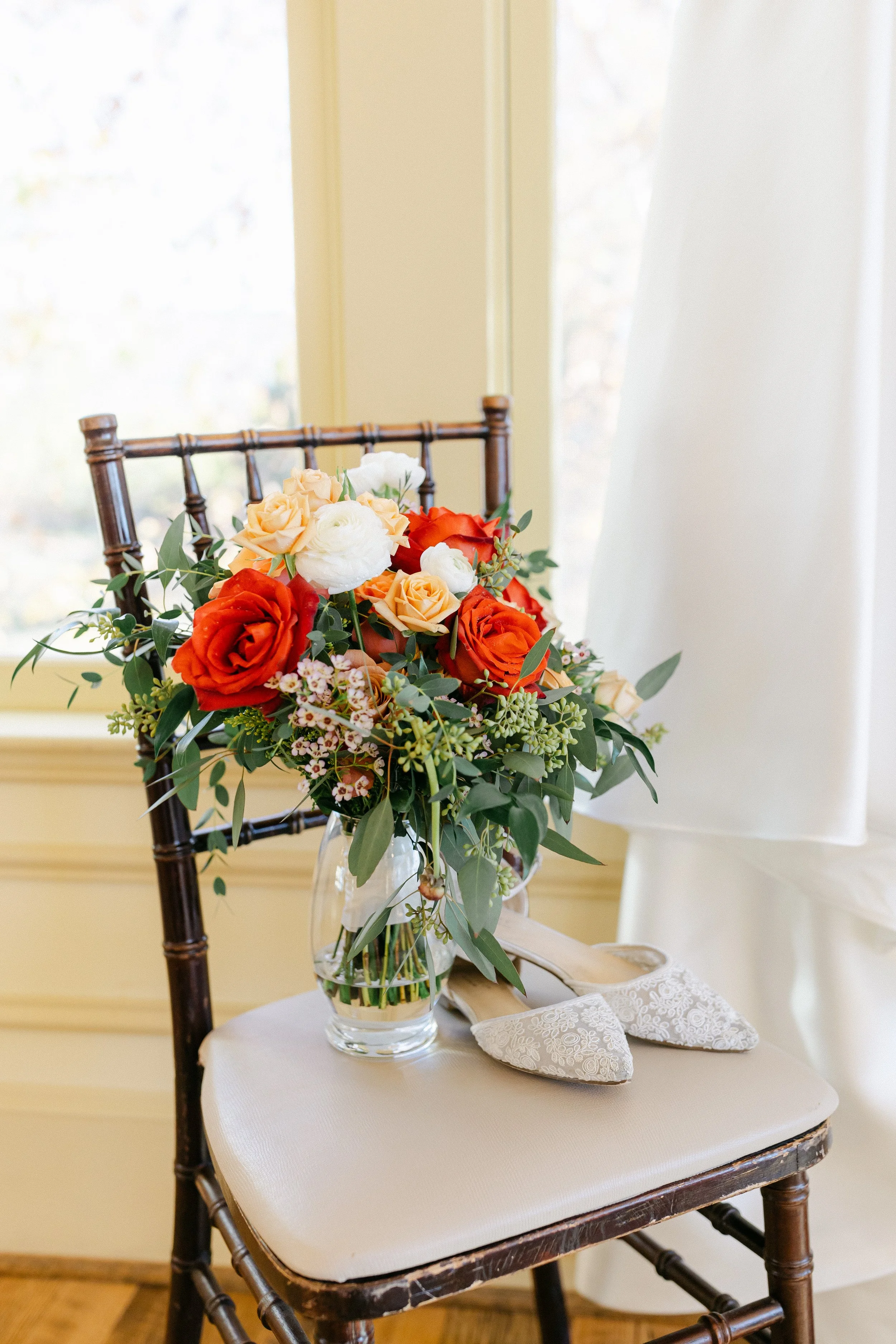 A bouquet of colorful flowers placed on a wooden chair with a pair of white embroidered shoes beside it, in a bright room with yellow walls and natural light.