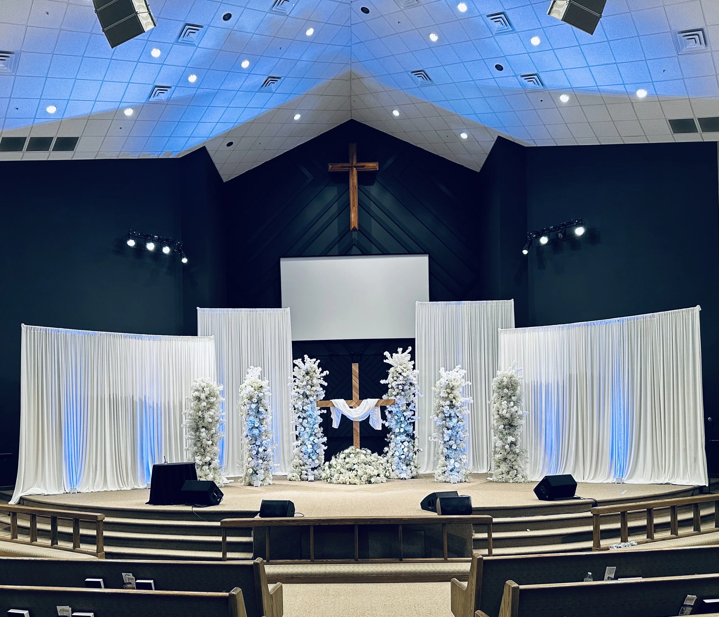 Empty stage in a church or chapel decorated for a wedding with a cross, white flowers, drapes, and lighting.