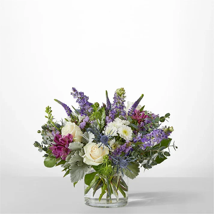 A colorful bouquet of flowers in a clear glass vase, including white roses, purple and pink flowers, and green foliage, set against a plain white background.