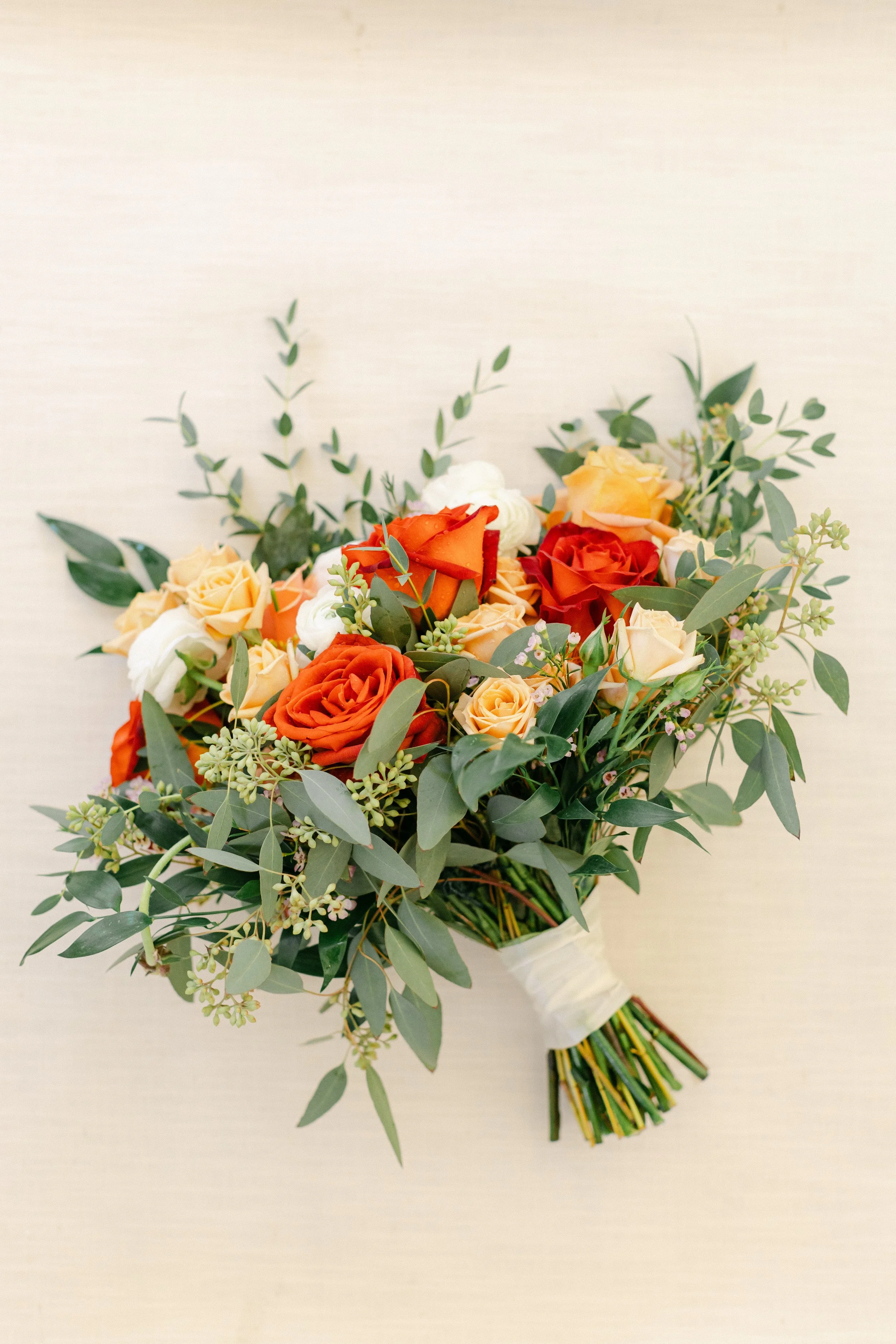 A colorful bouquet of orange, cream, and white roses with greenery on a neutral background.
