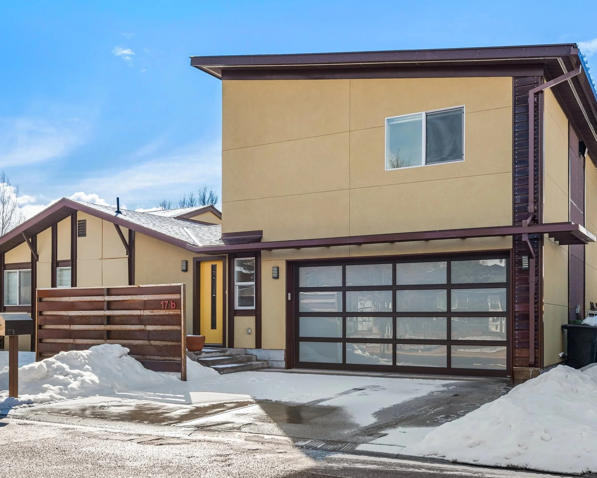 Modern two-story house in a snowy neighborhood with a yellow front door, large window, and a glass garage door, under a blue sky.