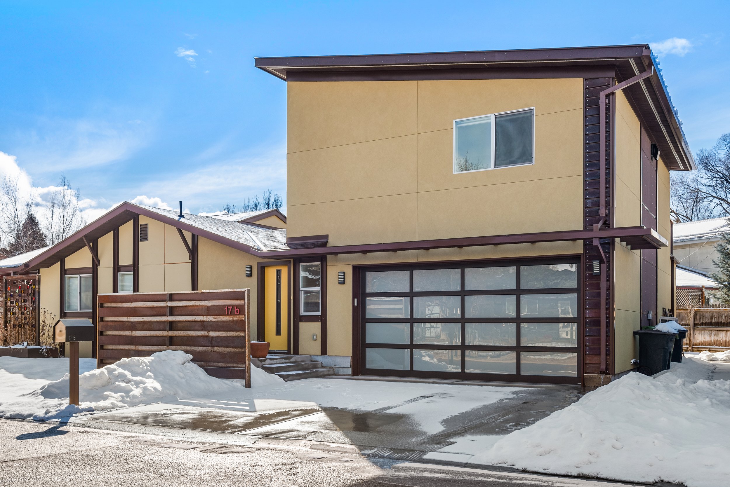 Modern two-story house with a yellow and brown exterior, large front garage with glass doors, snow on the ground, and a clear blue sky.