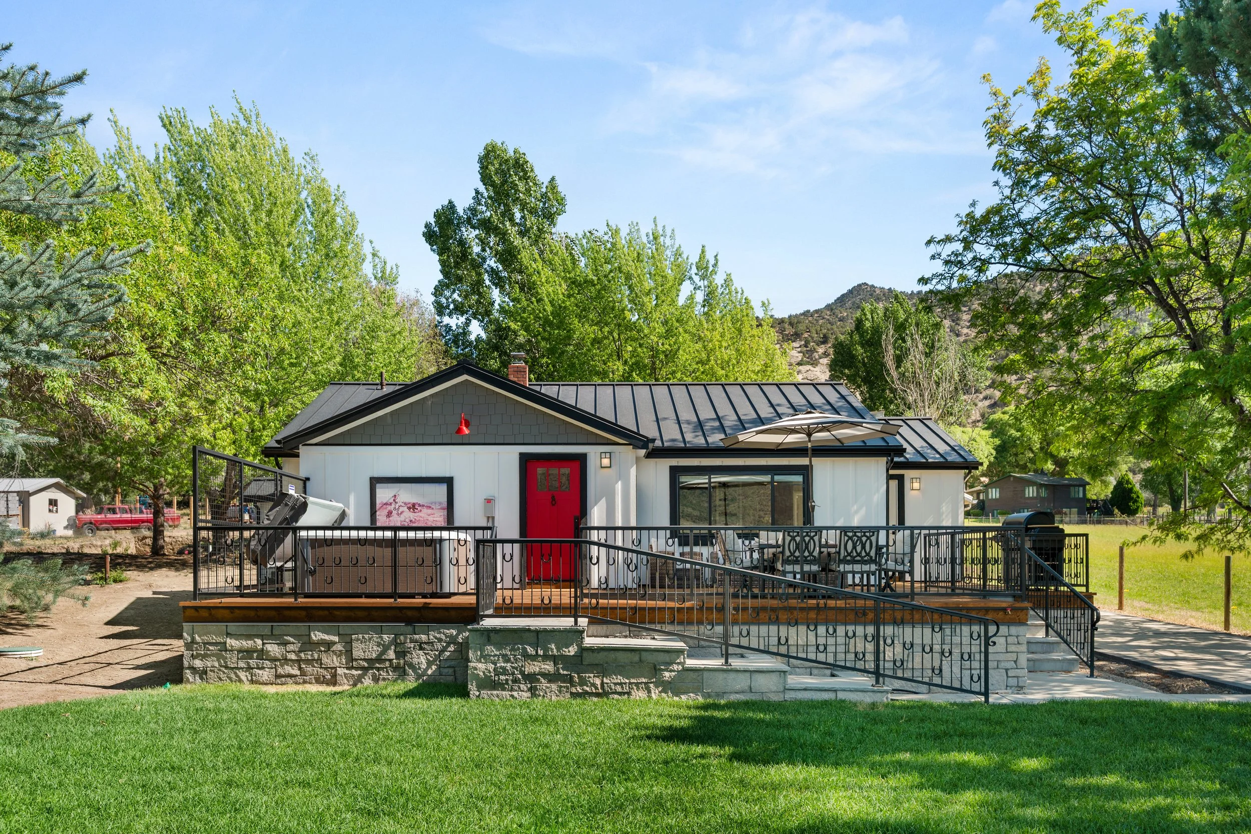 A house with a black metal roof, white exterior walls, and a red front door is surrounded by green trees and a grassy yard. There is a large deck with outdoor furniture and an umbrella, with stairs leading down to the yard.